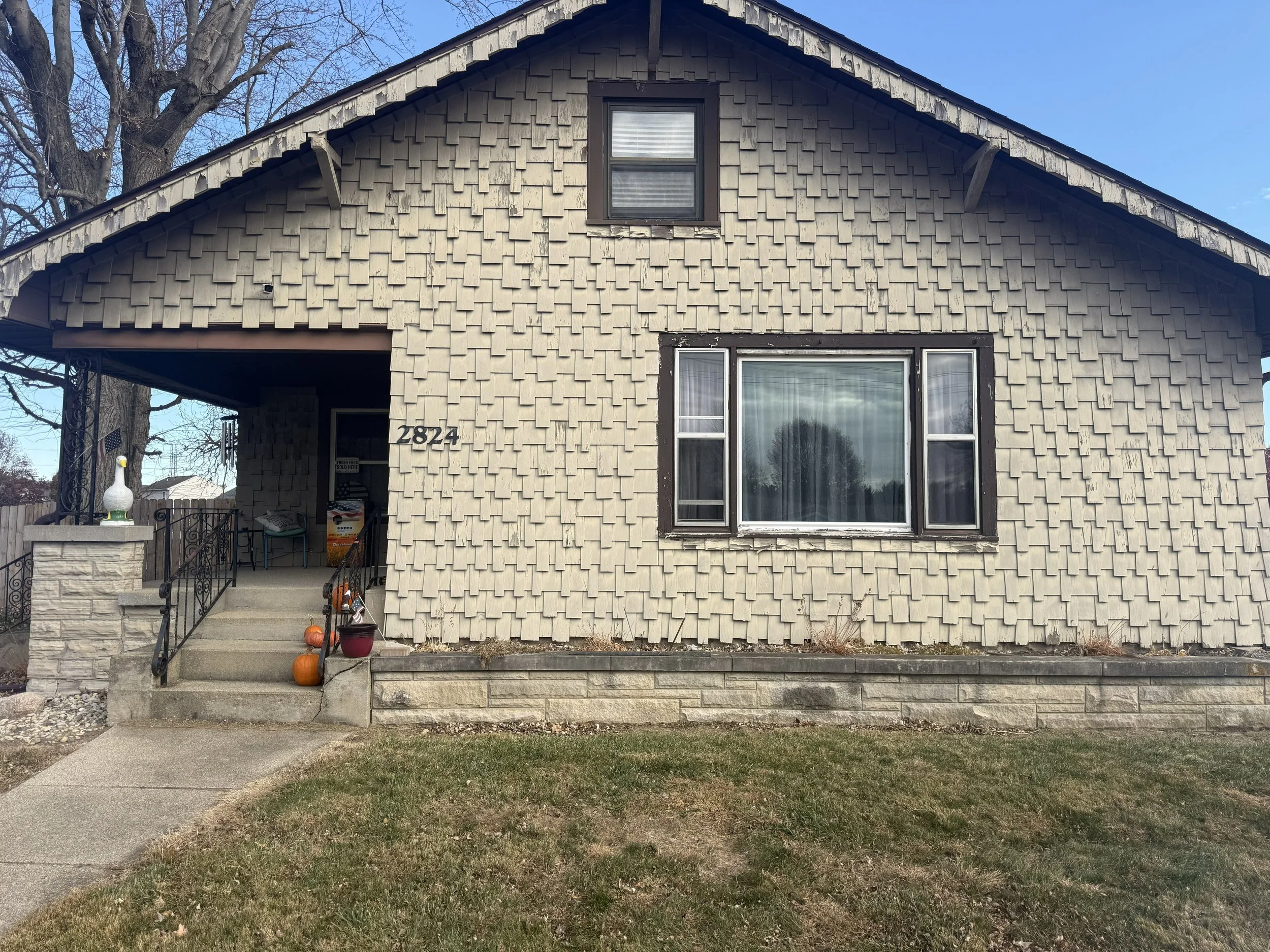 Front view of a house with beige shingle siding, a small porch with steps, pumpkins on the steps, and a large window. The house number 2824 is visible near the entrance.