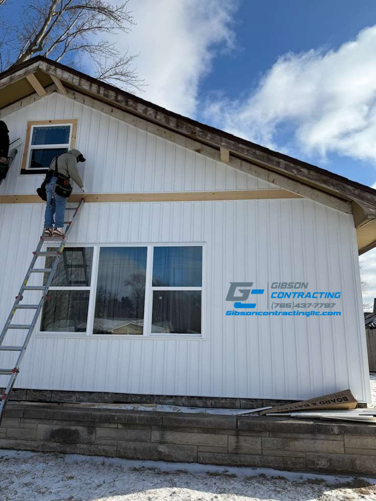 Person working on the exterior of a white house, standing on a ladder, installing trim or siding, with a logo of Gibson Contracting on the wall, under a partly cloudy sky.