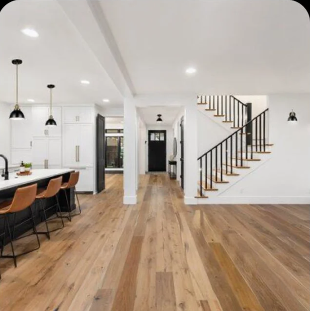 Interior of a modern home with a wooden floor, white walls, black staircase railing, kitchen area with a dining island and brown chairs, and multiple ceiling lights.