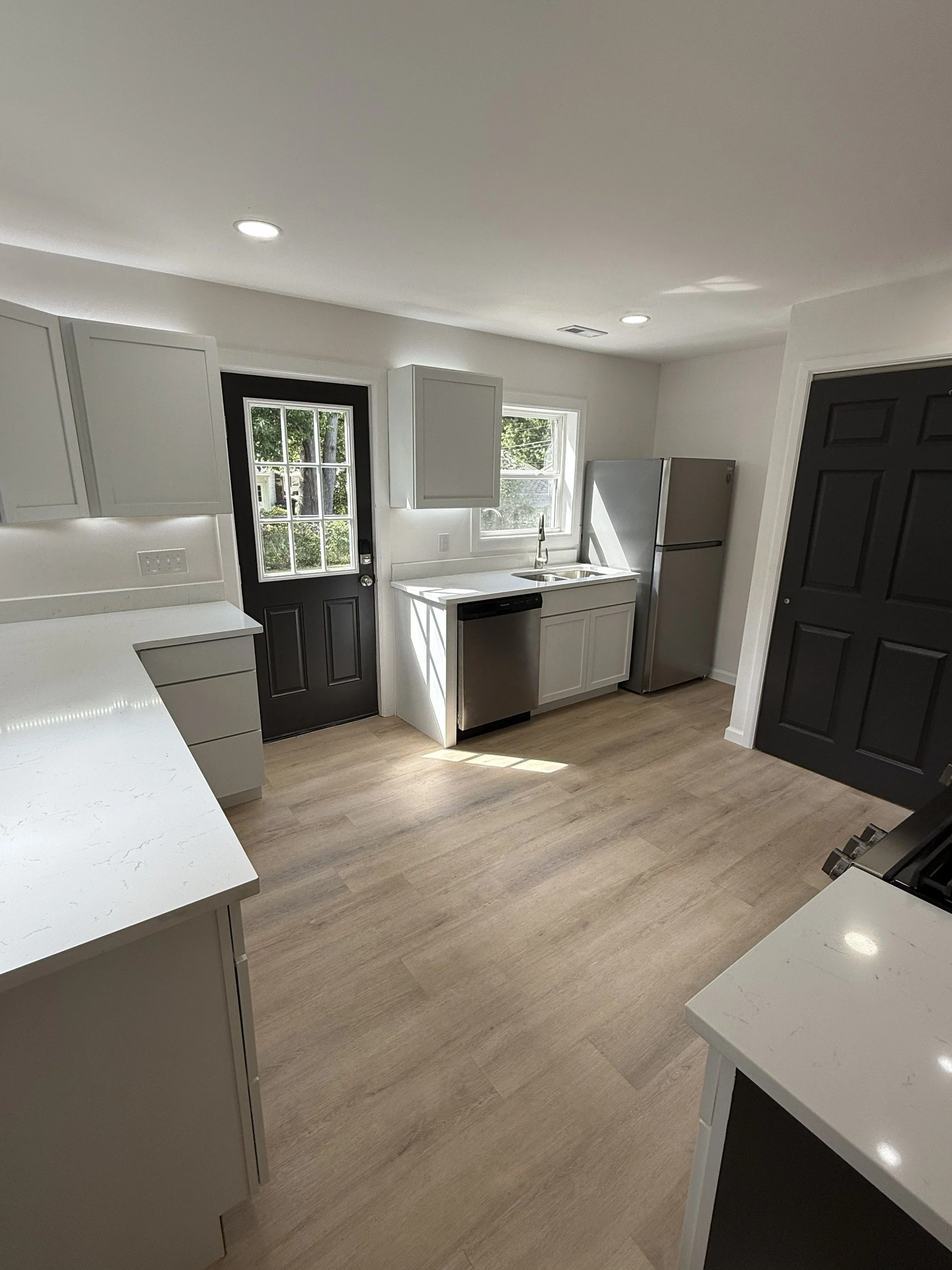 A modern kitchen with white countertops, white cabinets, a stainless steel refrigerator, a black door with a window, and a window above the sink. Light wood flooring and recessed ceiling lights are visible.