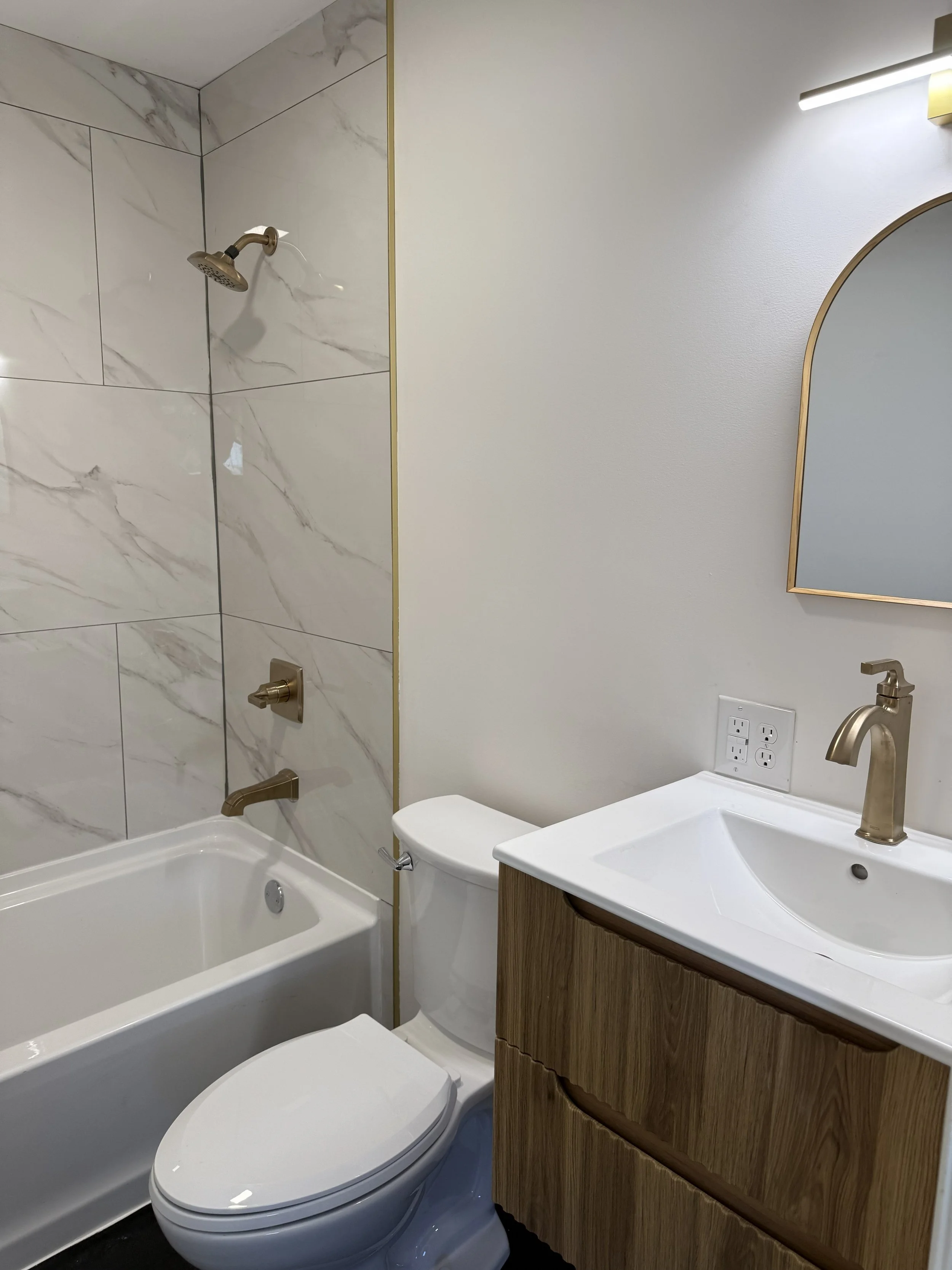 Bathroom with bathtub, toilet, small sink with wooden cabinet, mirror, lighting, and marble wall tiles.
