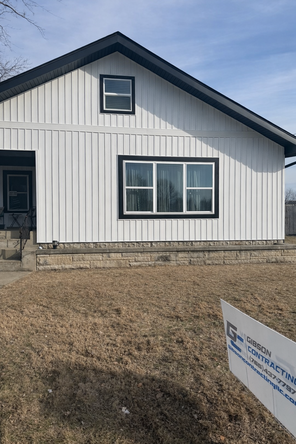 White house with black trim around windows and roof, concrete steps leading to front door, and a sign in the yard advertising a contracting company.