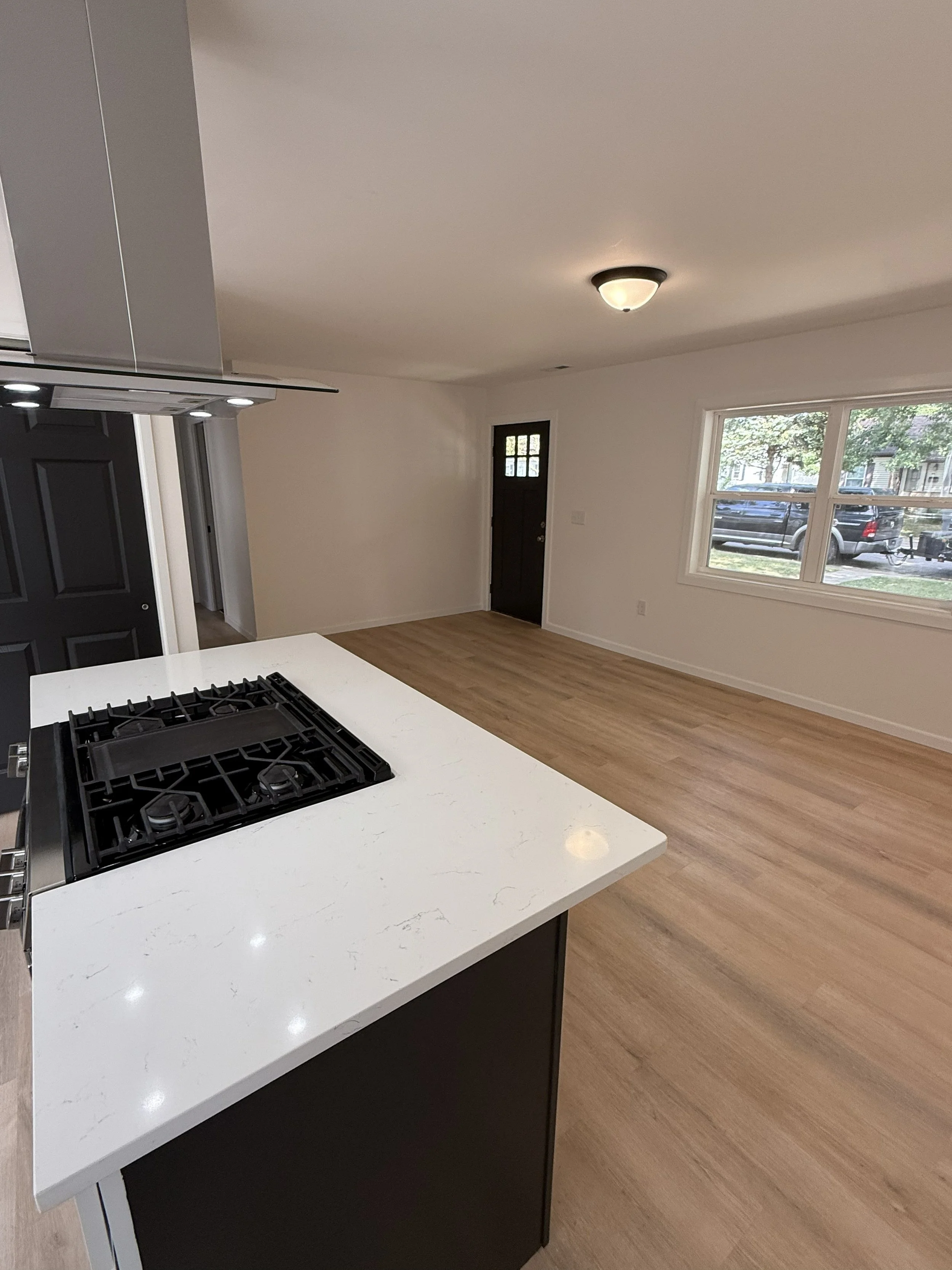 Empty living room and kitchen area with a stove on a white countertop, large window showing a parked truck outside, and a black front door.