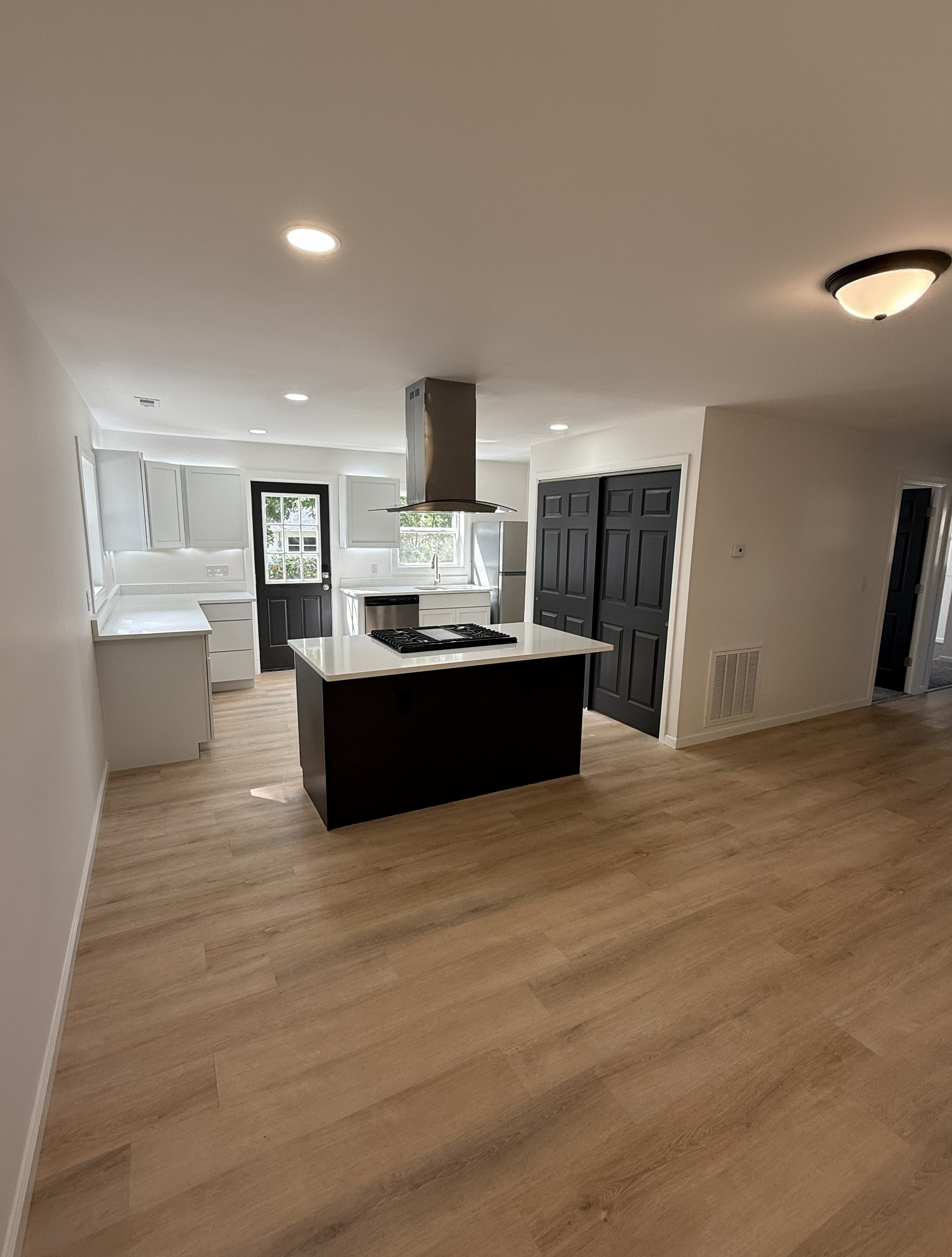 Modern kitchen with light wood flooring, white cabinets, black accents, and stainless steel appliances including an island with a gas stove and a range hood.