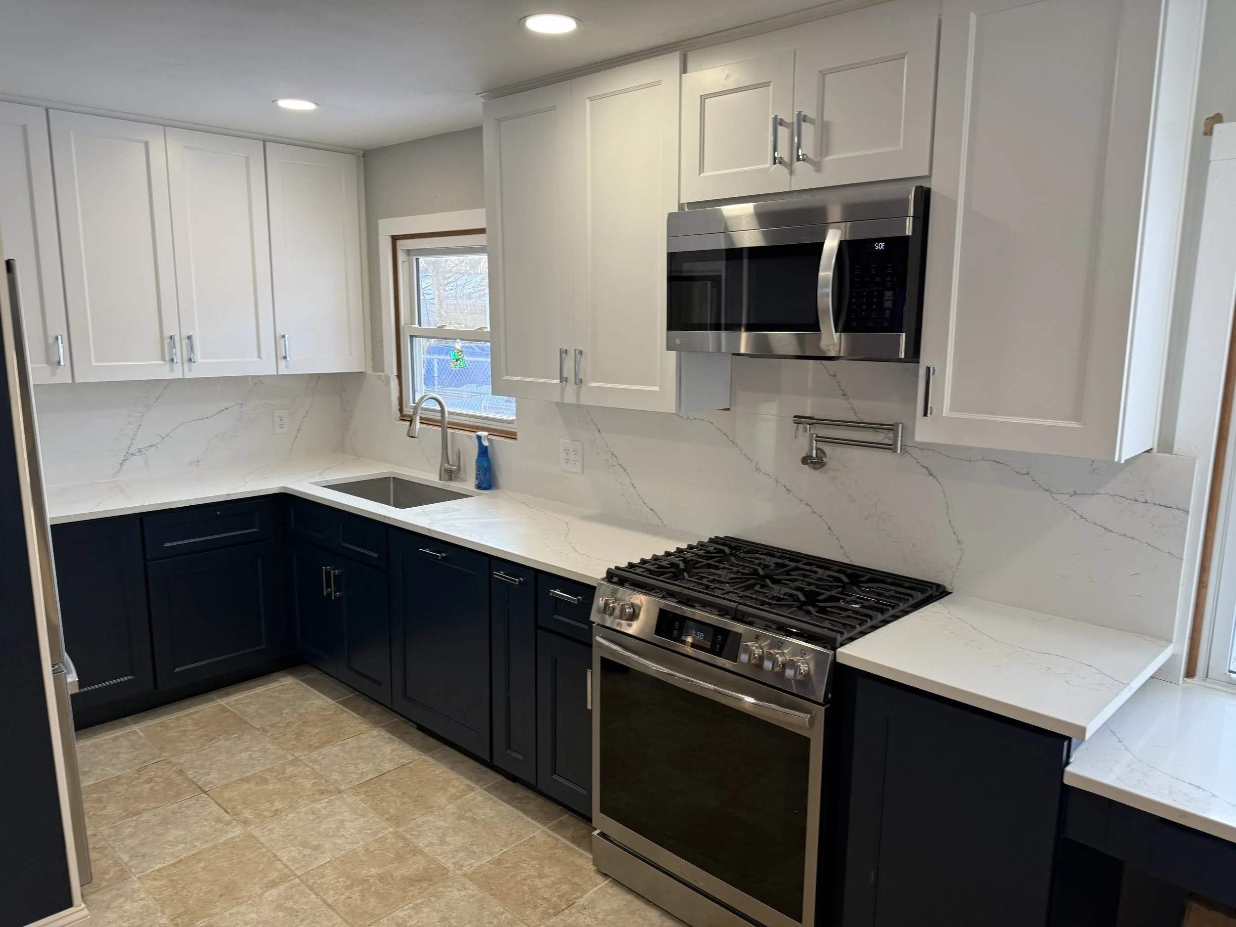 Modern kitchen with white upper cabinets, dark lower cabinets, marble countertop and backsplash, stainless steel microwave, oven, and sink, with a window above the sink and beige tiled floor.