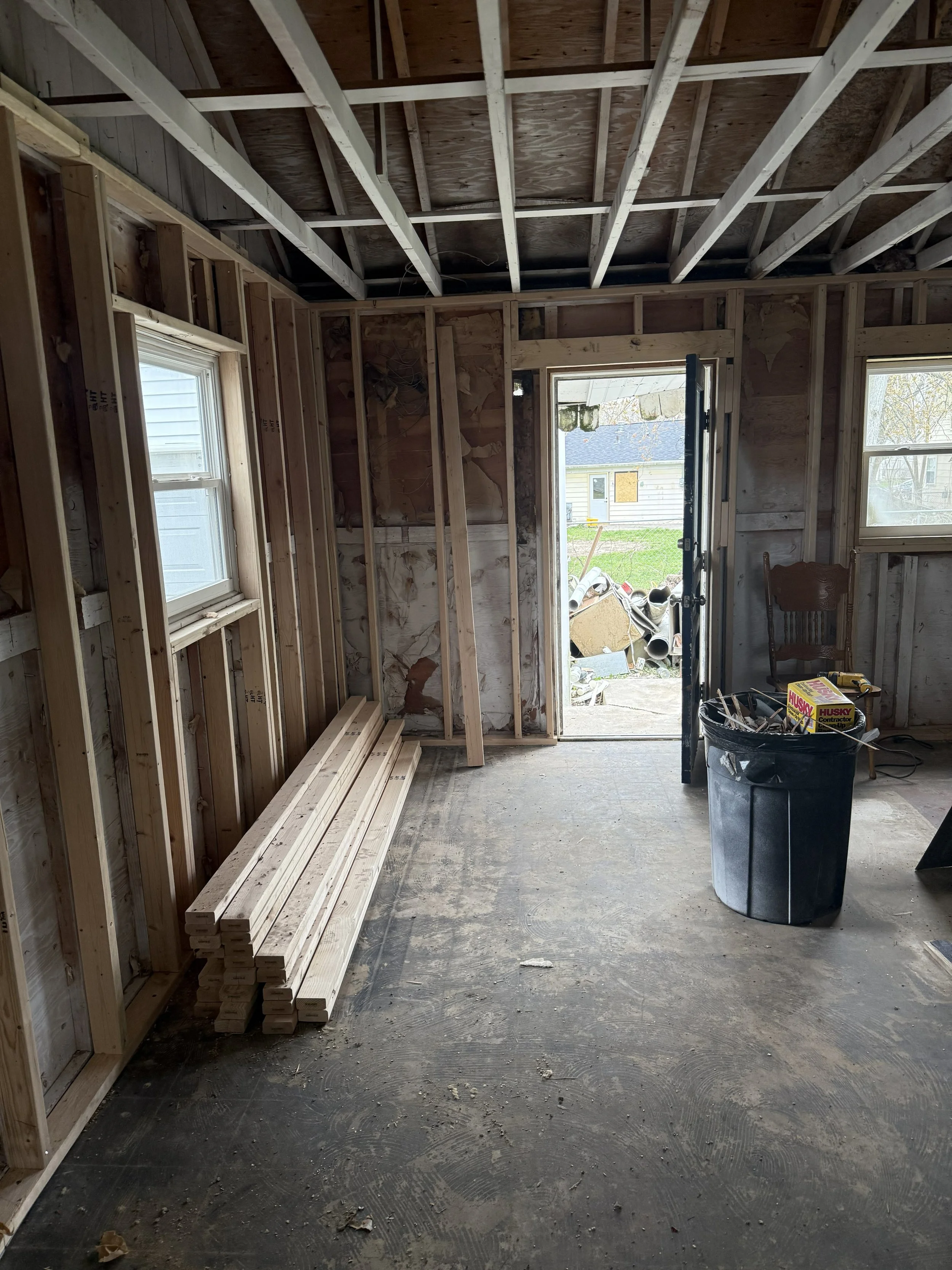 Interior of a house under construction with exposed wooden framing, windows, and a door opening leading outside, construction materials including lumber and a trash bin, and a chair in the background.
