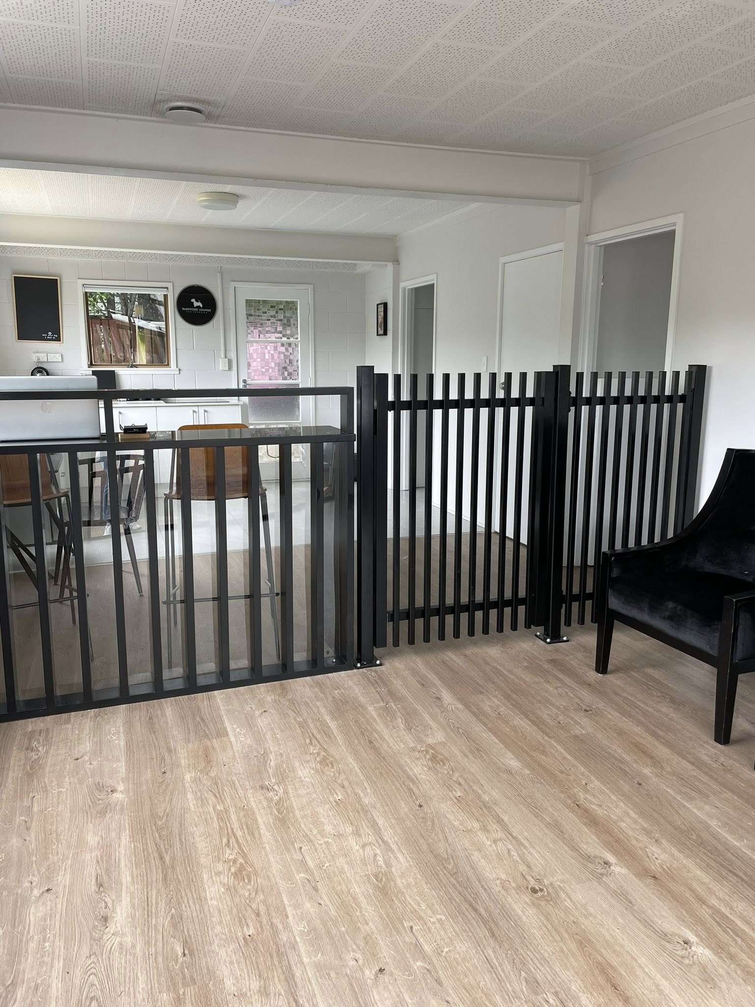 Interior of a room with a black pet gate, a wooden chair, a black chair, and a glass door leading outside; white walls and ceiling with a wood floor.