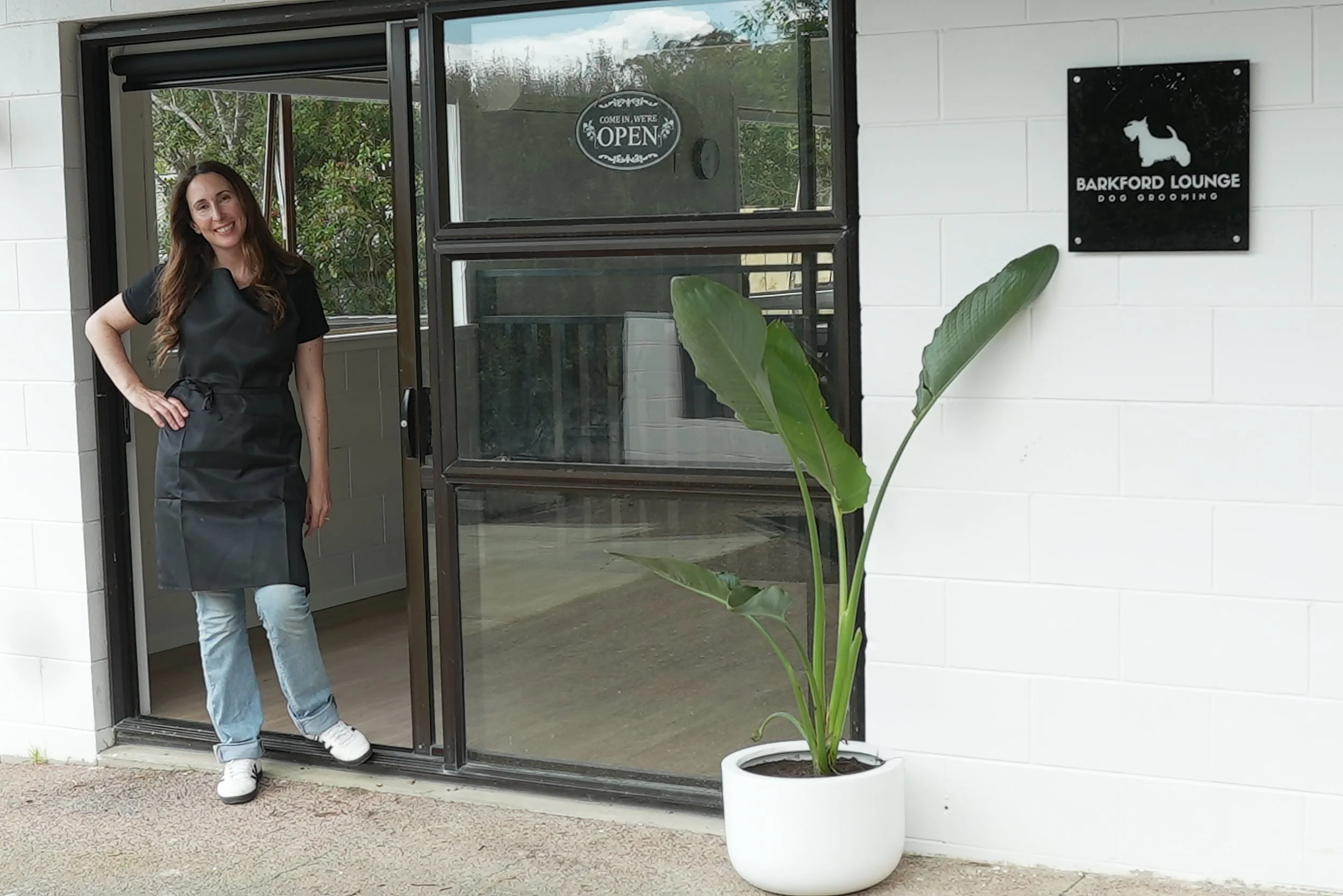 A woman standing at the entrance of Barkford Lounge, a dog grooming salon, wearing a black apron and jeans, smiling. There is a large potted plant with green leaves beside her. The salon's sign is mounted on the white wall to her right, and a window with an
