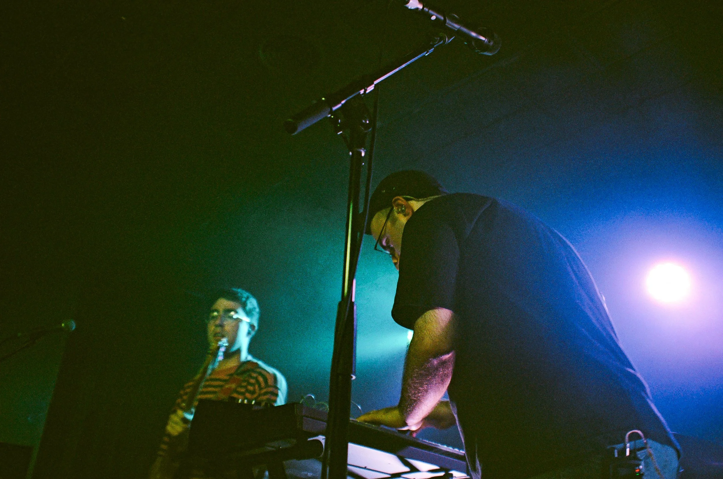 Two men are performing a music set on stage in a dimly lit environment with colorful lighting. One man is wearing glasses and a striped shirt, while the other man is wearing a black shirt and glasses, leaning over equipment.