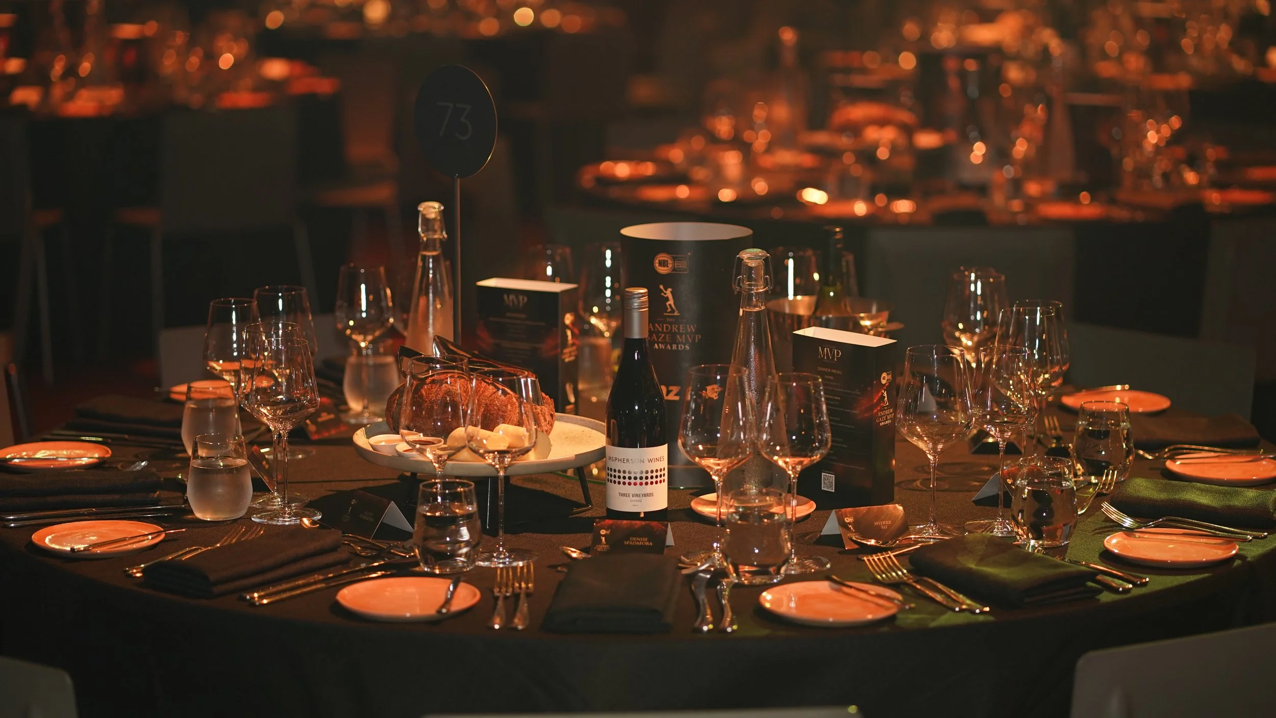 An elegantly set dining table at a formal event with multiple wine glasses, a bottle of wine, a plate of bread, and various menus and place cards, under dim warm lighting.