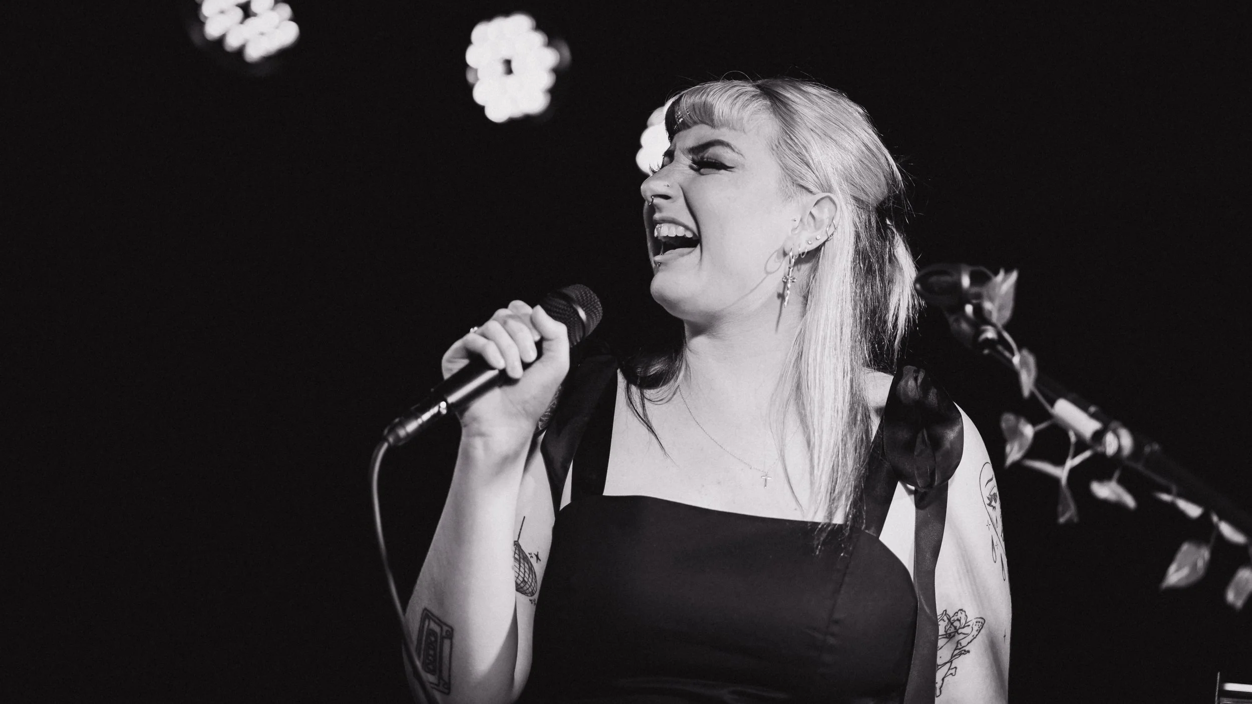 A woman singing passionately into a microphone on stage, with a dark background and some blurred decorative lights.