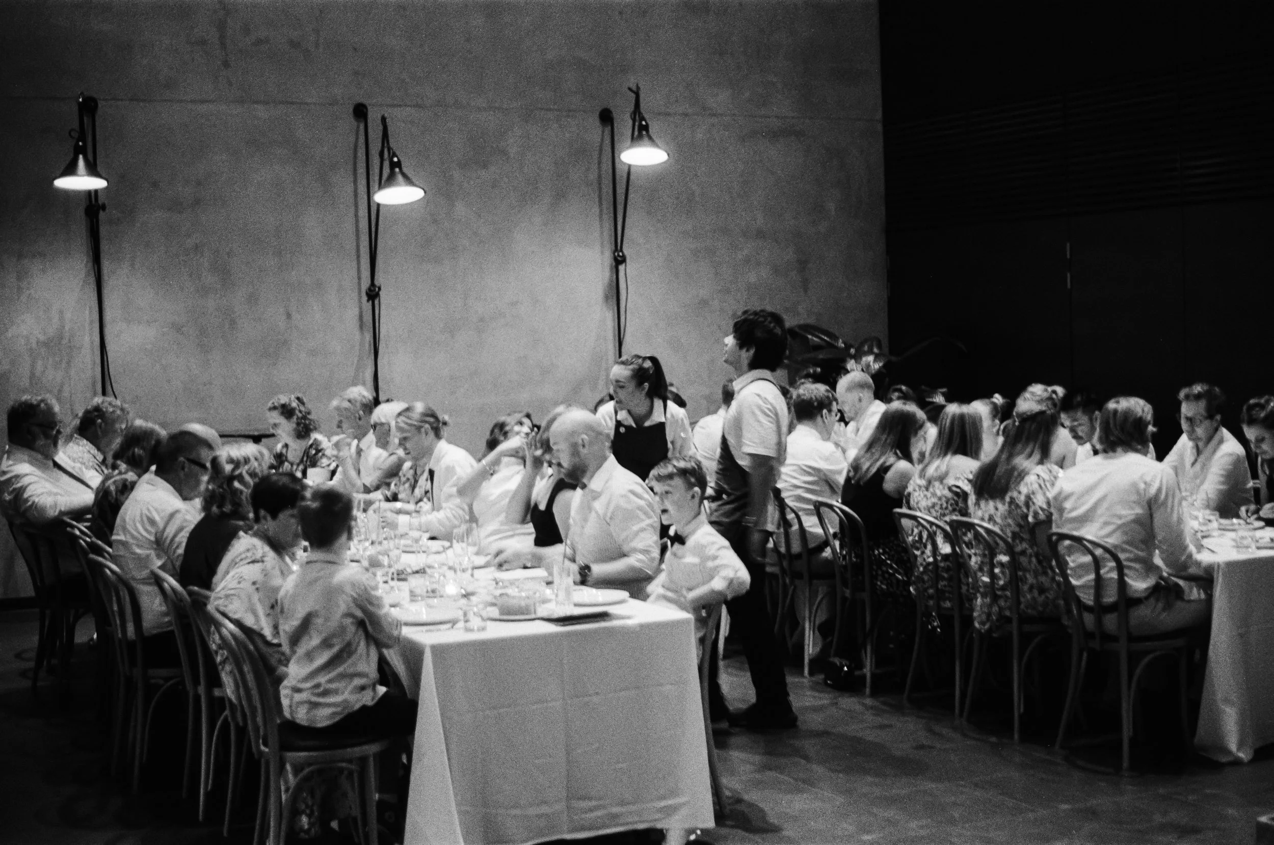 A black-and-white photo showing a large dining gathering in a modern, minimalist restaurant or banquet hall. People are seated at long tables covered with white tablecloths, engaged in conversation and dining. Several waitstaff are serving food to gu