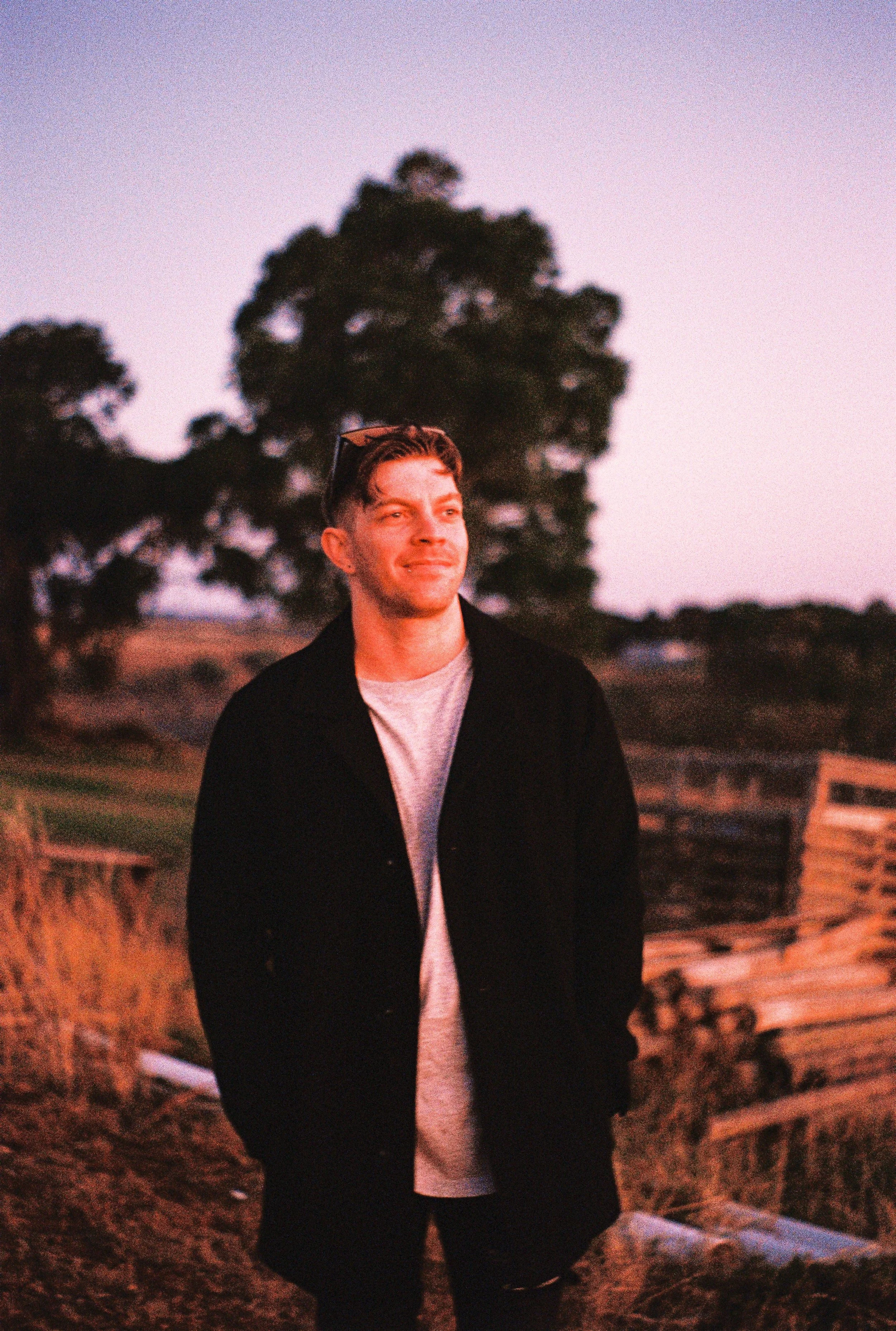 A young man with short hair and sunglasses on his head, smiling, outdoors during sunset, with trees and wooden fences in the background.