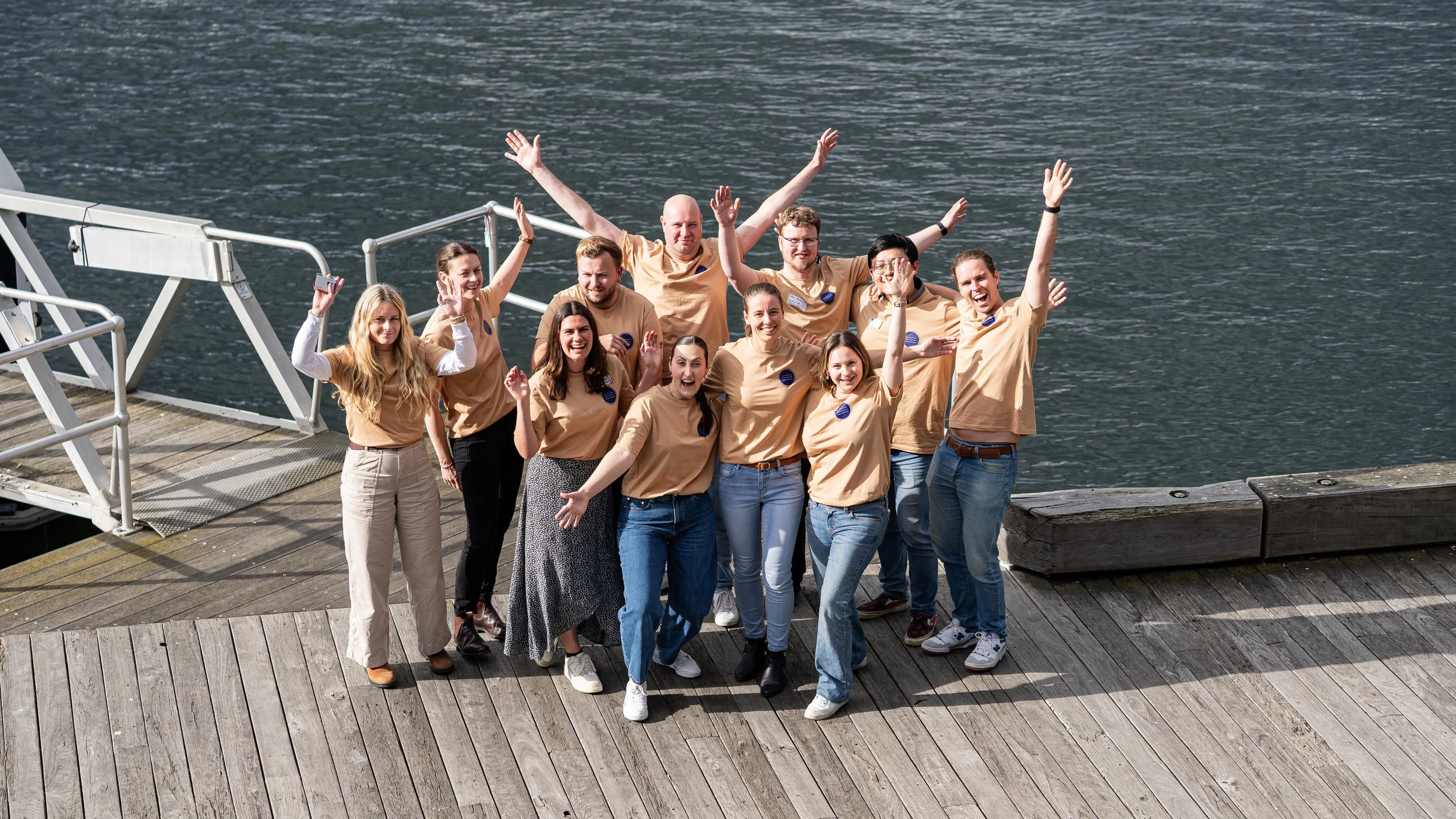 Group of people smiling and celebrating on a wooden dock by the water, with some raising their hands and one taking a selfie.