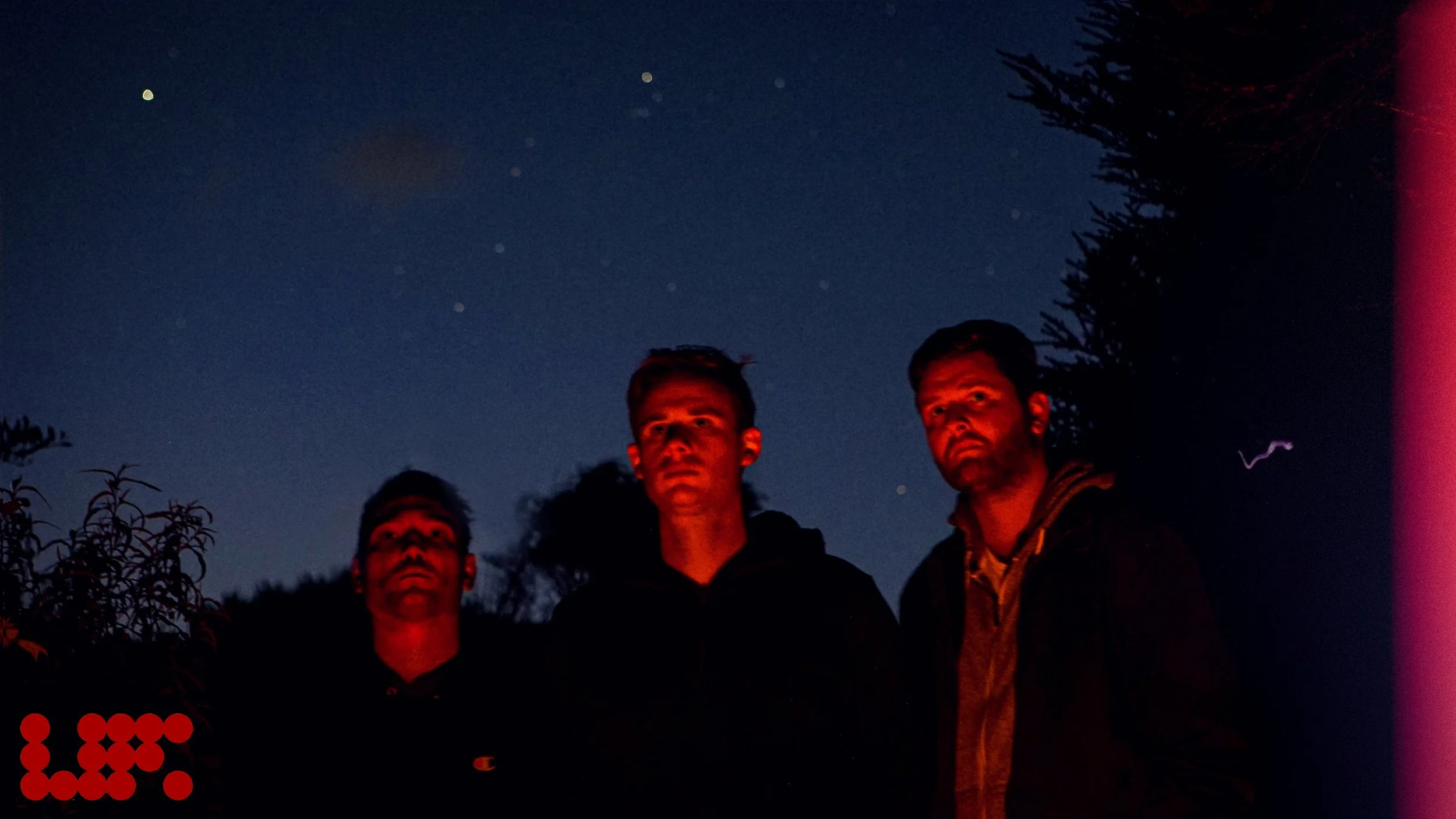 Three men standing outdoors at night under a starry sky, with trees around them, illuminated by a red light.