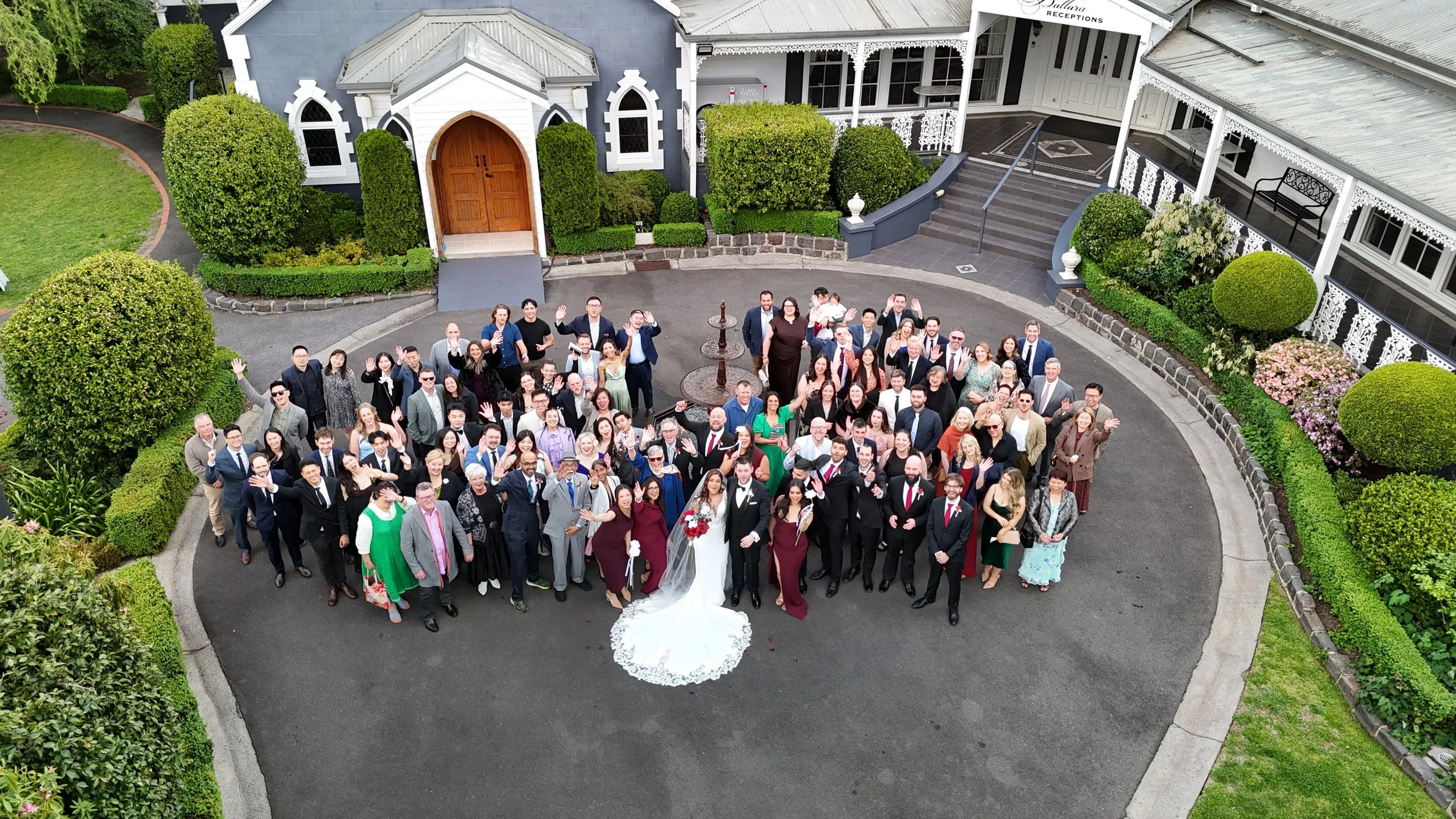 A large group of people gathered outdoors in front of a house, celebrating a wedding. The bride and groom are near the center, with the bride in a white wedding gown and the groom in a suit. The group is smiling and waving at the camera, standing on 