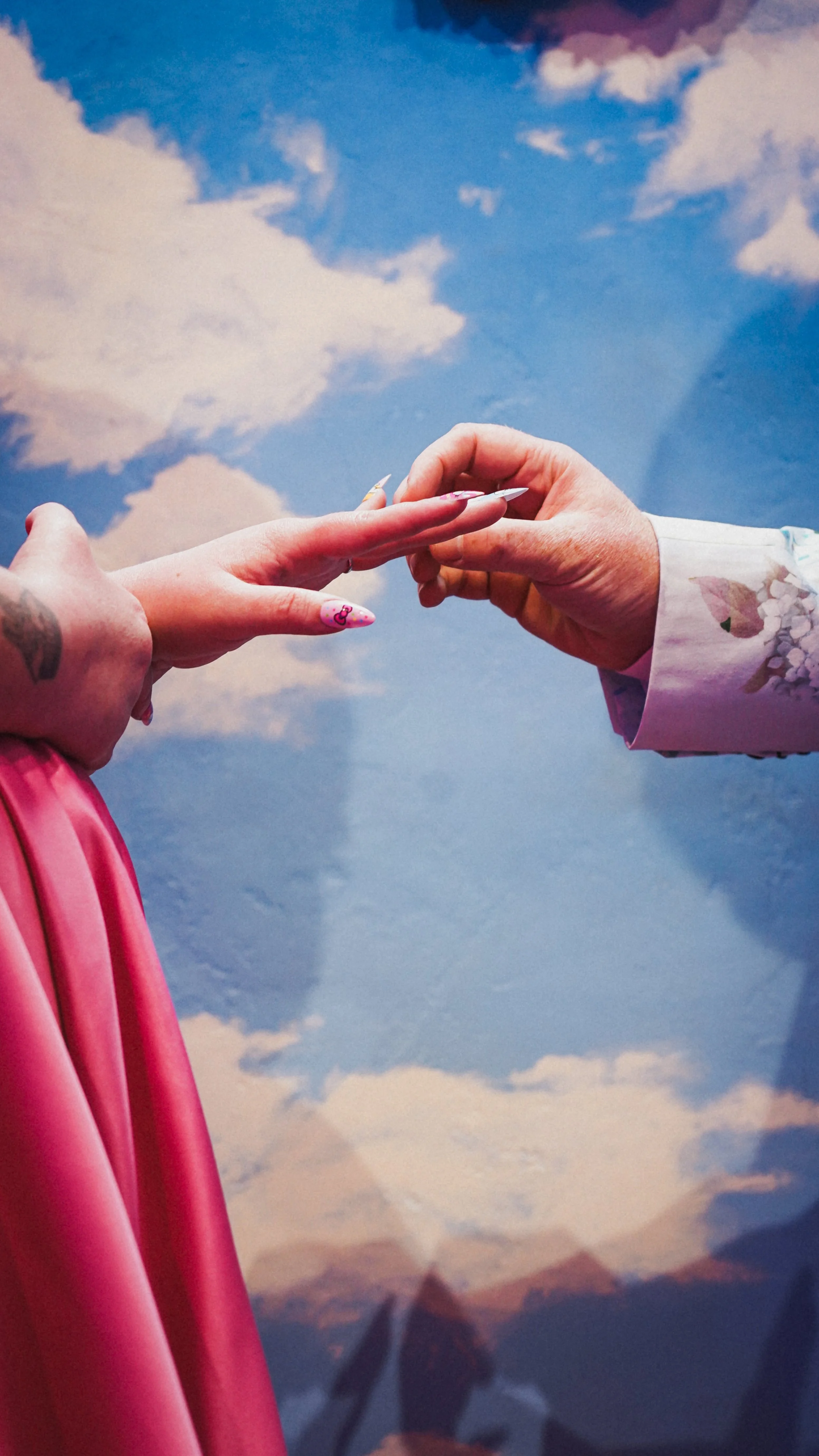 A person with long nails reaching out to hold another person's hand, against a backdrop of the sky with clouds and mountains.