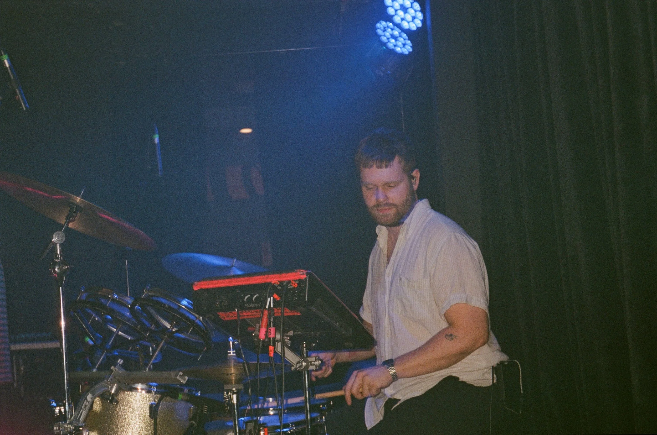 A man with a beard and mustache playing electronic music on a synthesizer or keyboard in a dark, dimly lit setting, possibly a concert or club, with blue stage lighting and black curtains in the background.