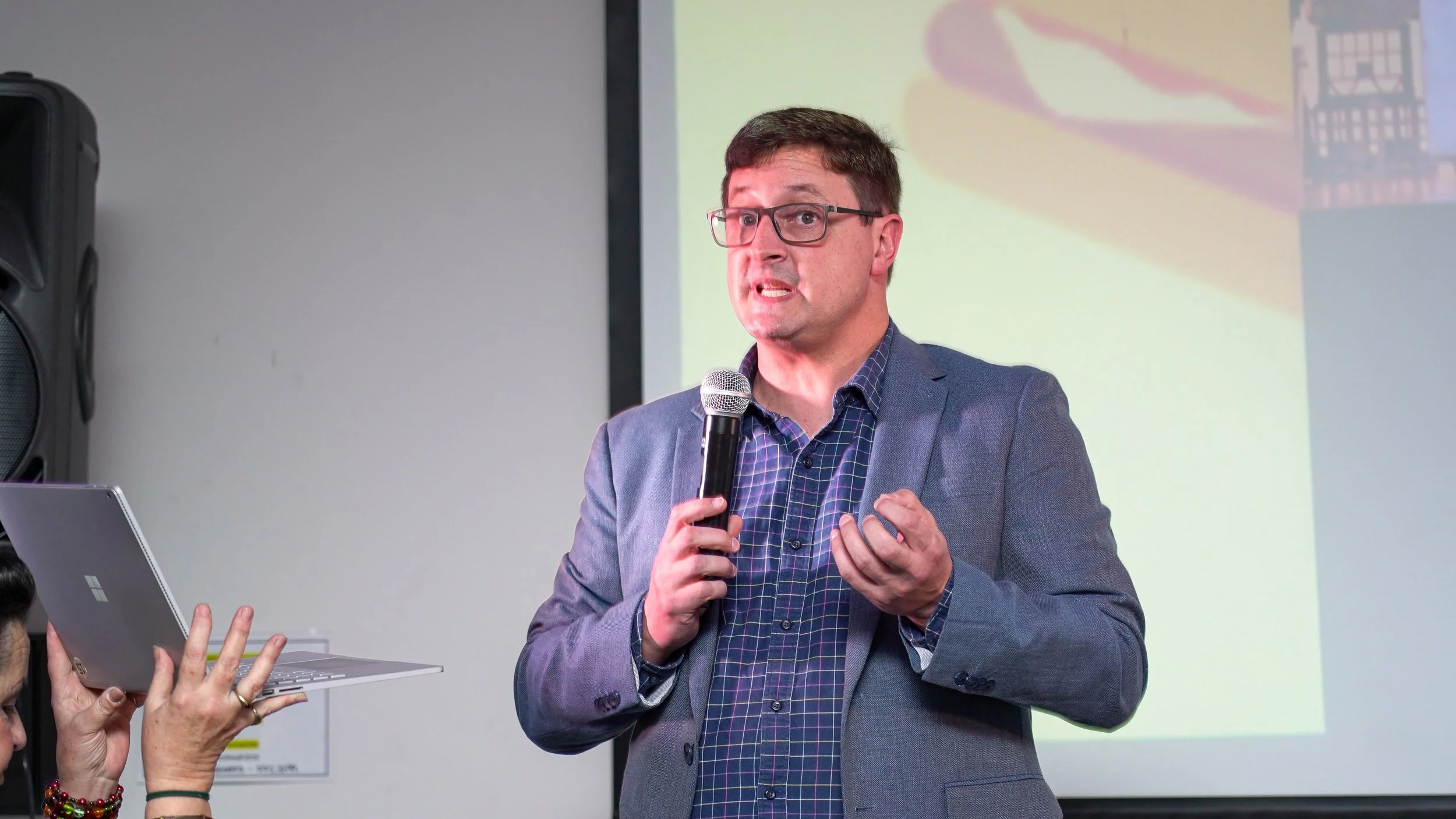 A man with glasses, wearing a blazer and checkered shirt, speaking into a microphone during a presentation or speech in a room with a projector screen in the background.