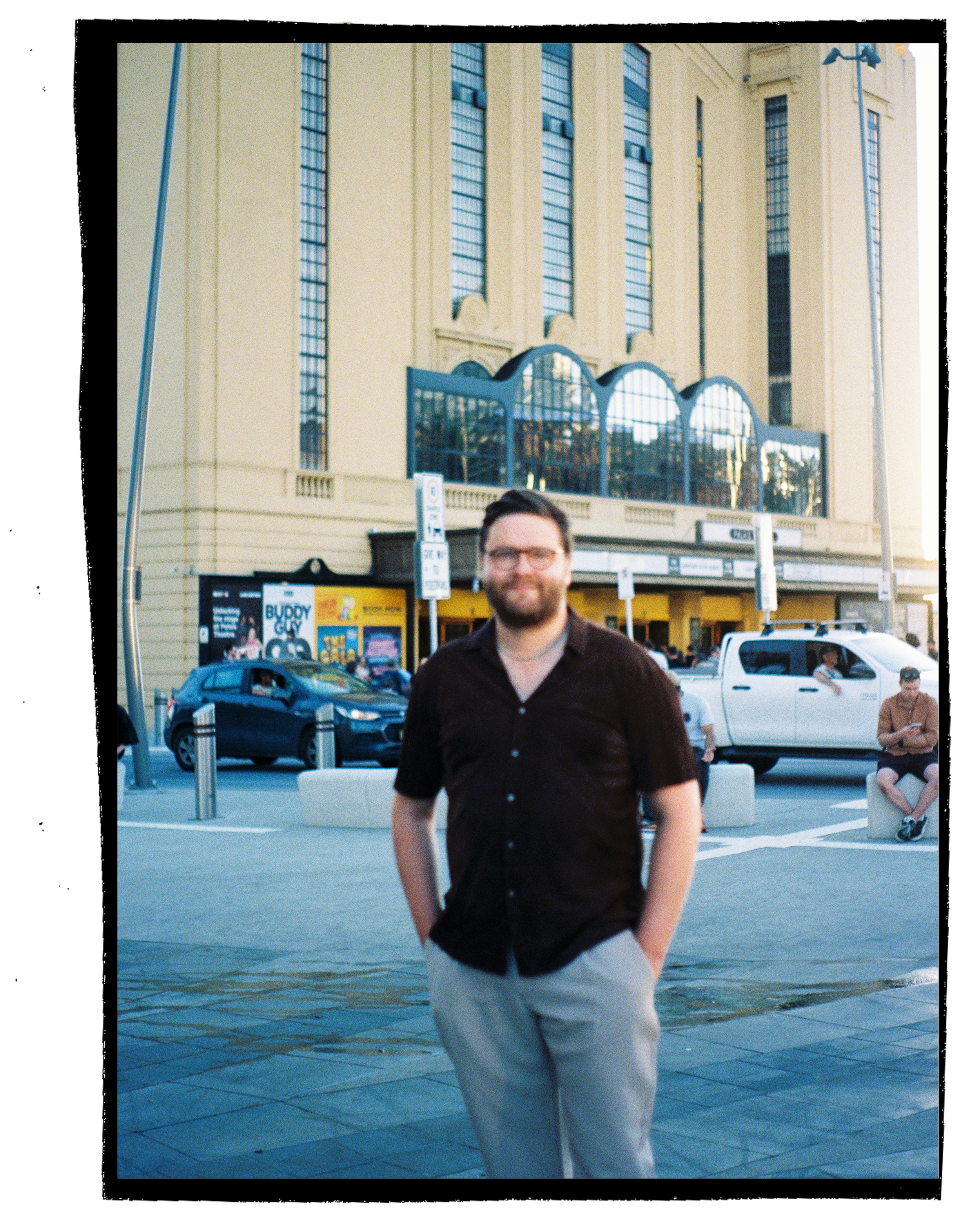 A man with glasses and beard walking along a waterfront sidewalk with water in the foreground, people and cars in the background, and a large building with vertical windows behind him.