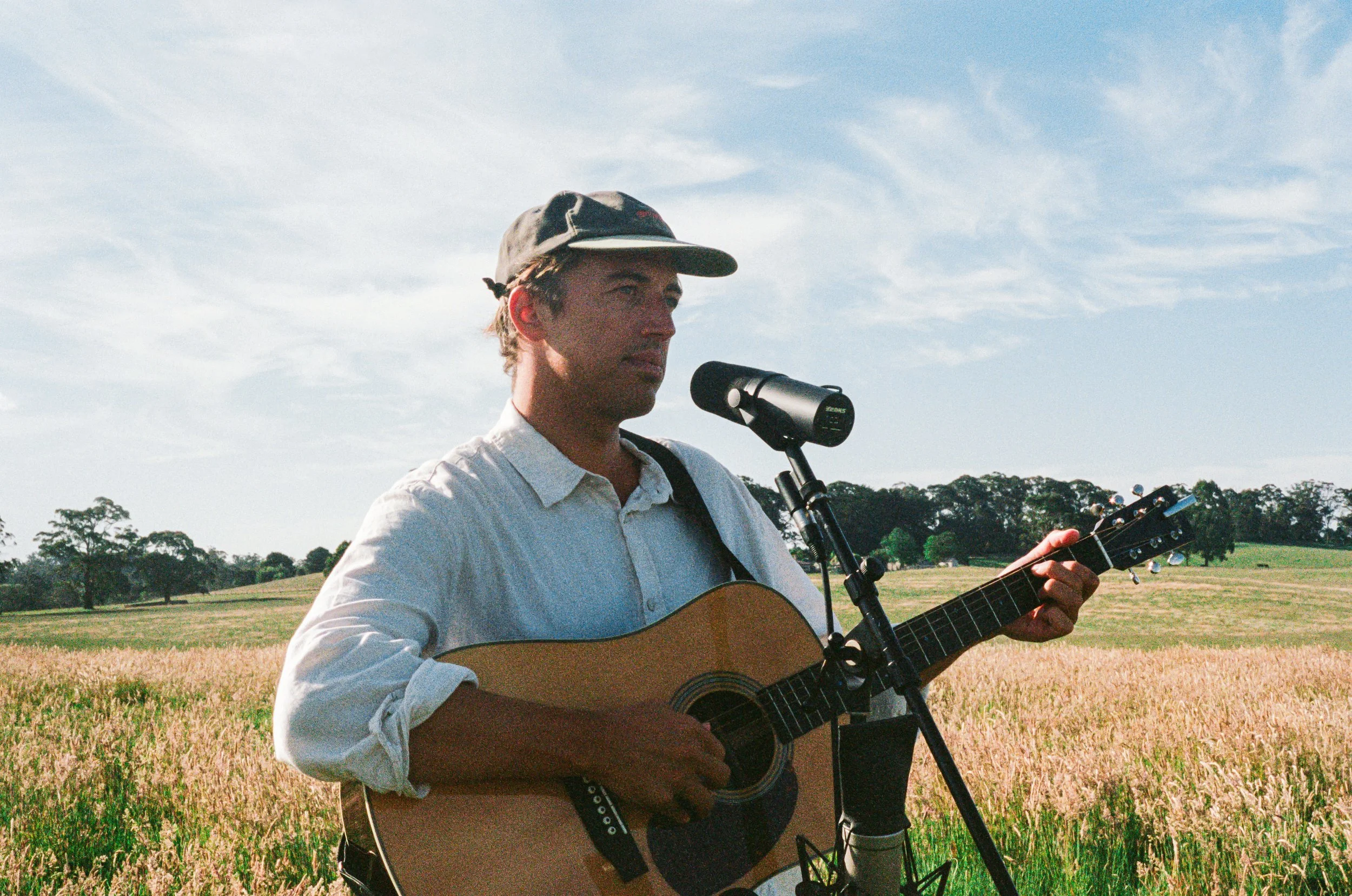 A man playing an acoustic guitar outdoors in a field under a partly cloudy sky, with trees in the distance.