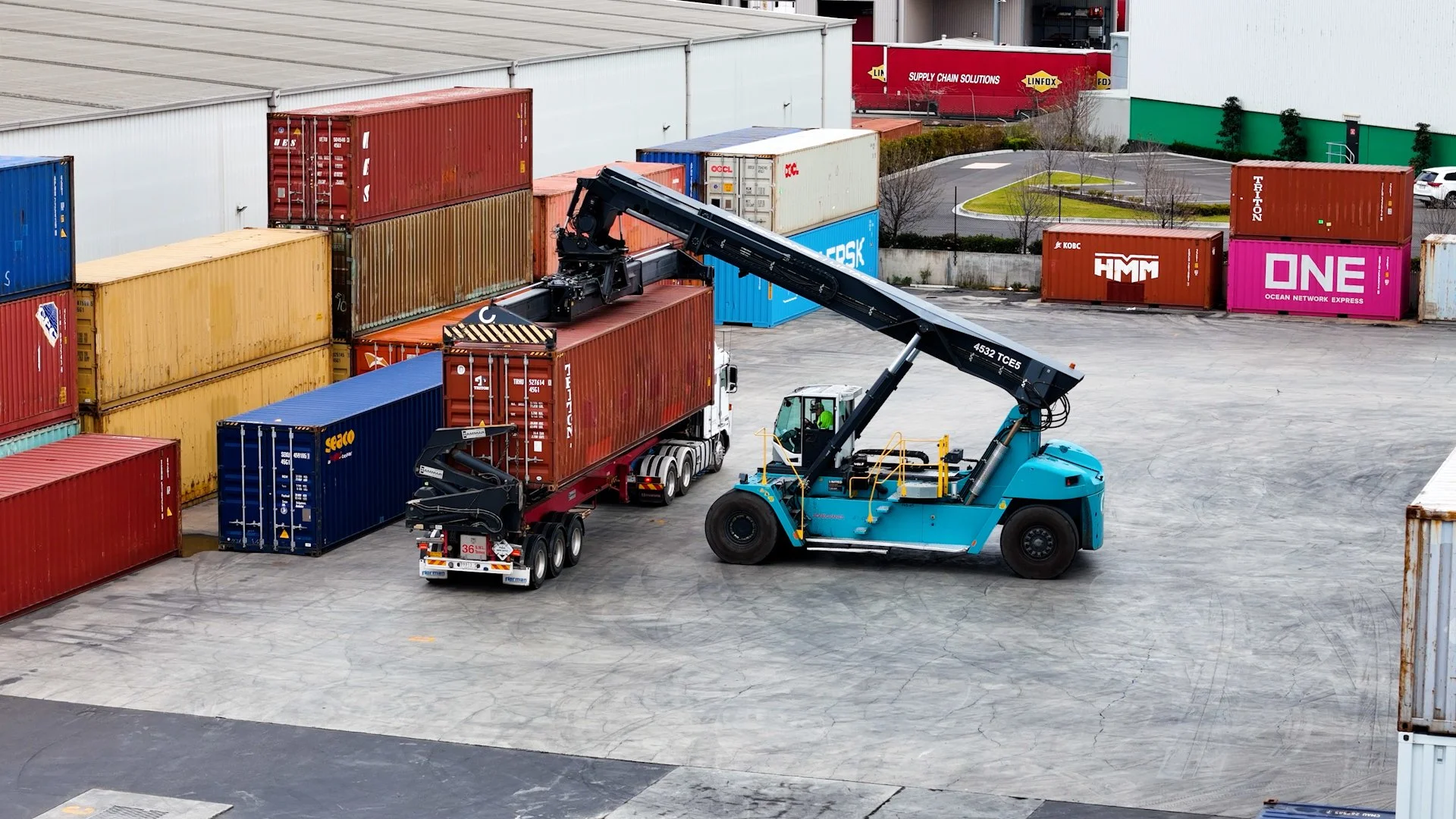 A forklift loading or moving a red shipping container at a cargo yard with various colored shipping containers stacked around.