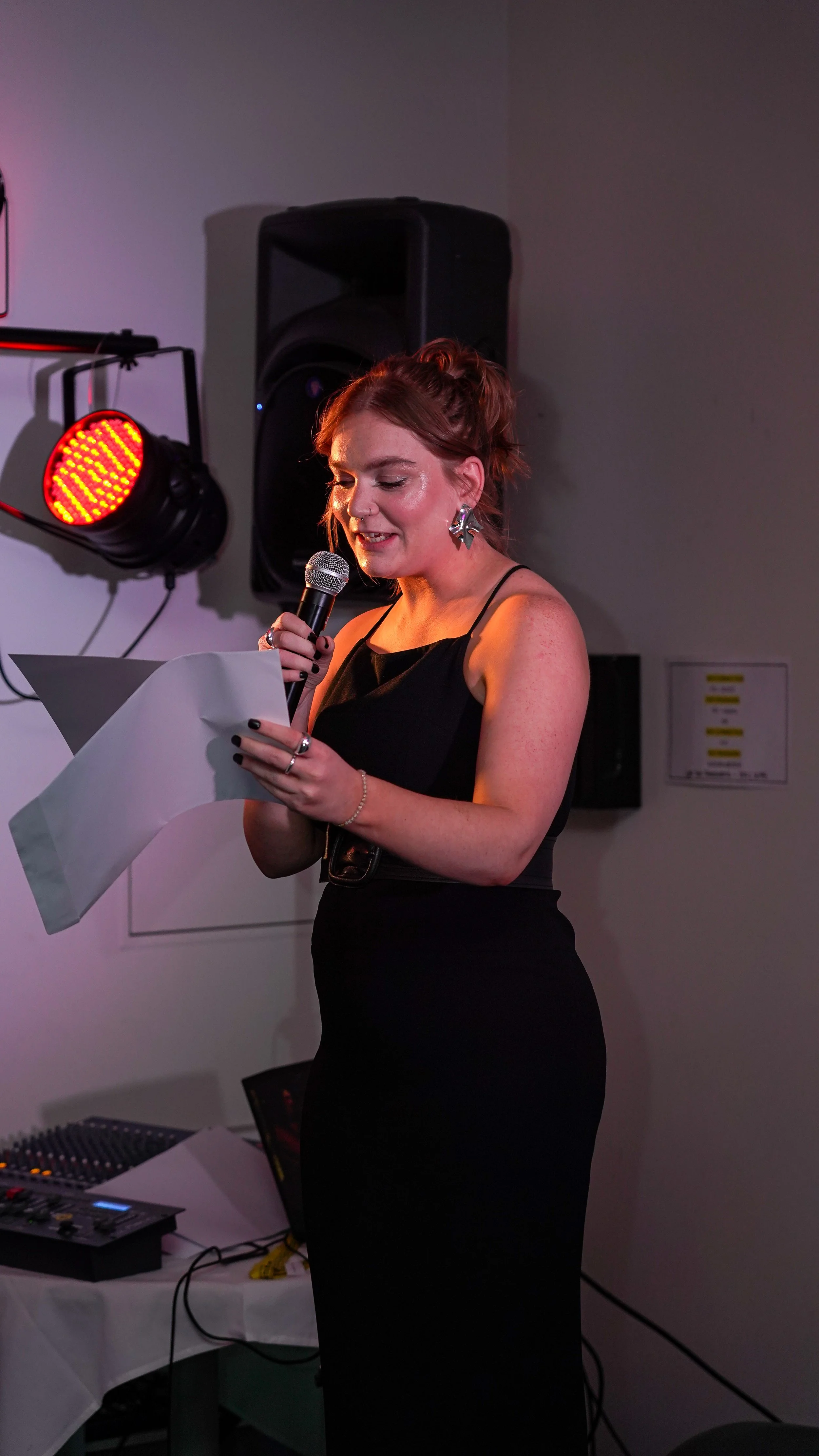 A woman in a black dress with earrings reading from paper and holding a microphone at an indoor event.