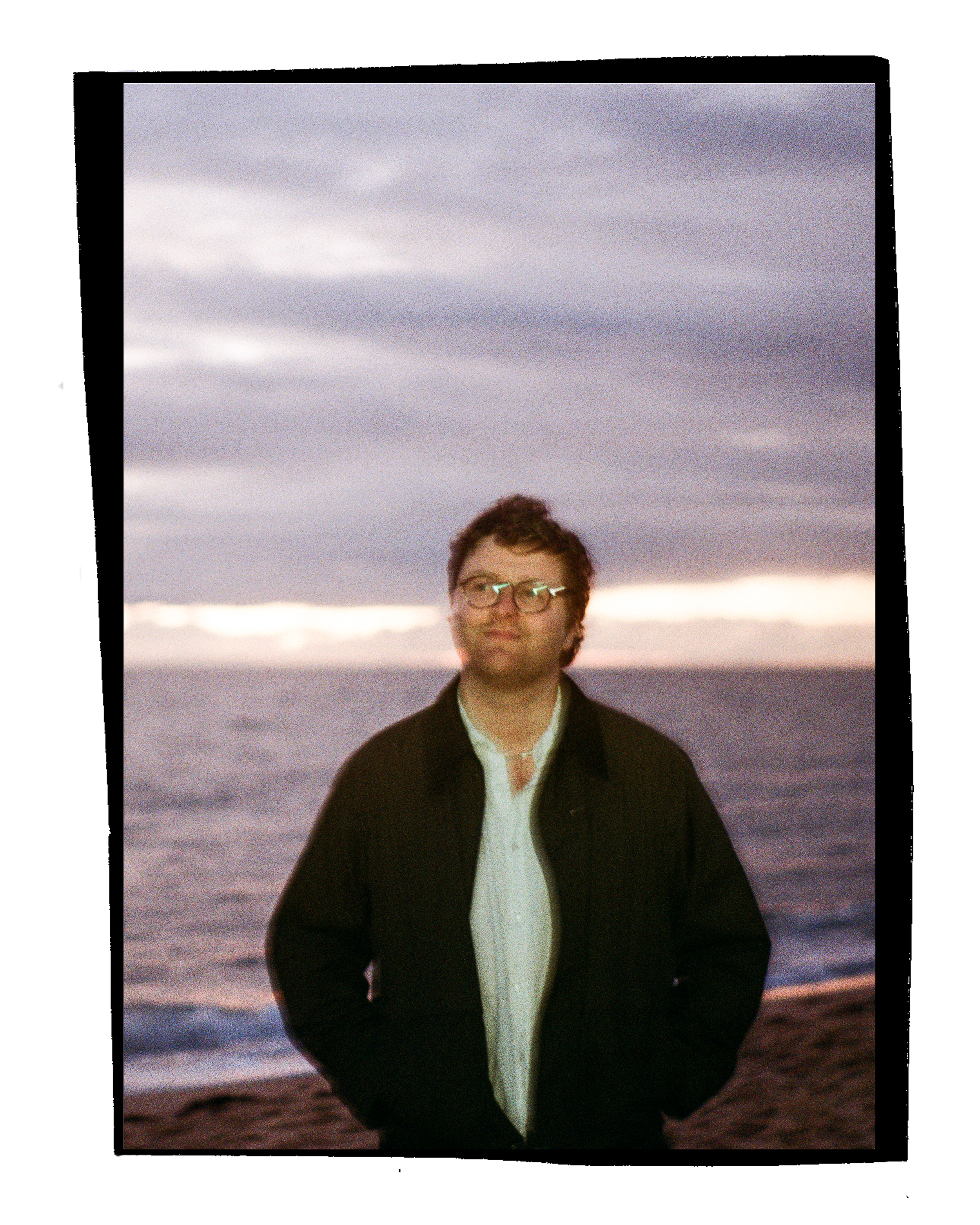 A man wearing glasses and a black jacket stands outdoors at sunset, partially smiling, with the ocean and cloudy sky in the background.