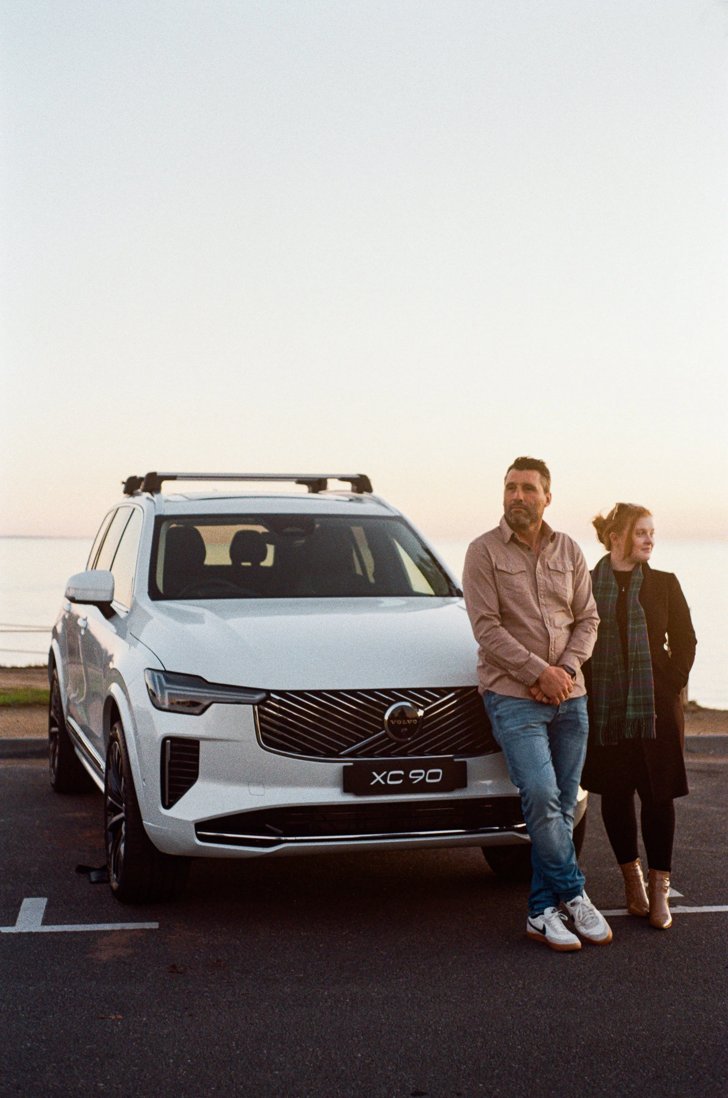 A man and a woman stand next to a white Volvo XC90 SUV parked on a paved area near a body of water during sunset. The man has dark hair and a beard, wearing a beige jacket and blue jeans, with his arms crossed. The woman has long hair tied back, wear