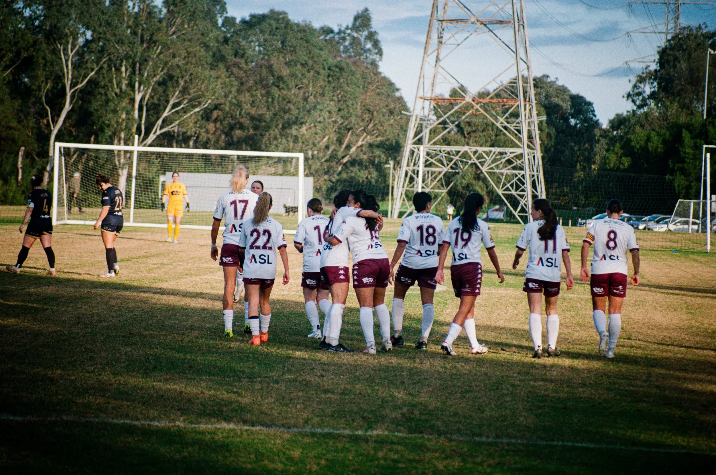 A group of women soccer players walking off the field, some hugging, with a goalpost and a soccer goalkeeper in yellow in the background, surrounded by trees and electrical towers.
