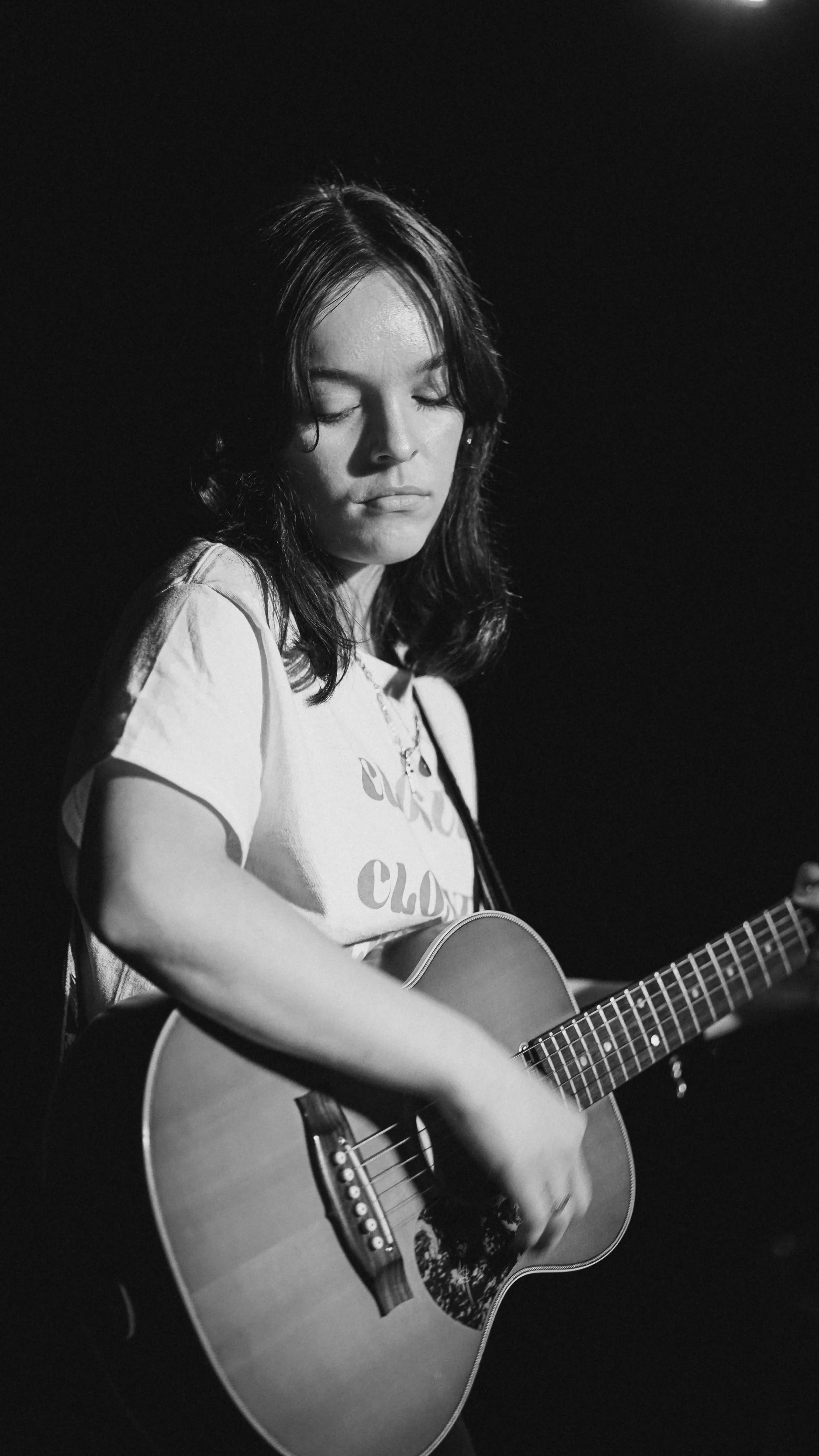 A young woman with dark hair playing an acoustic guitar on stage, eyes closed, in black and white.