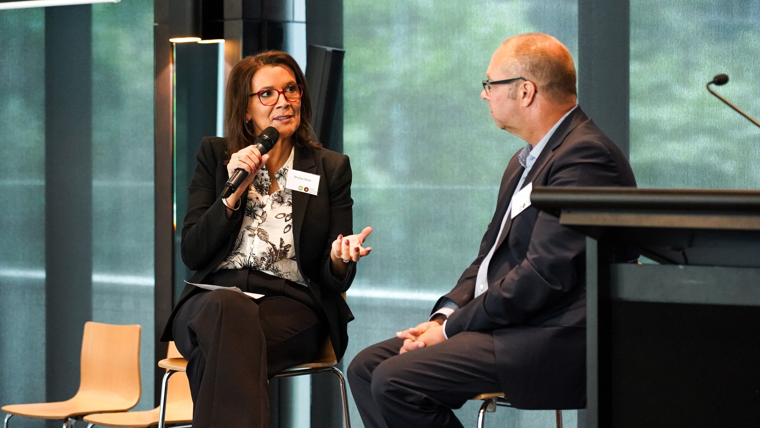A woman with red glasses holding a microphone and speaking to a man with glasses, who is sitting on a chair. They appear to be engaged in a discussion or interview in a professional setting.