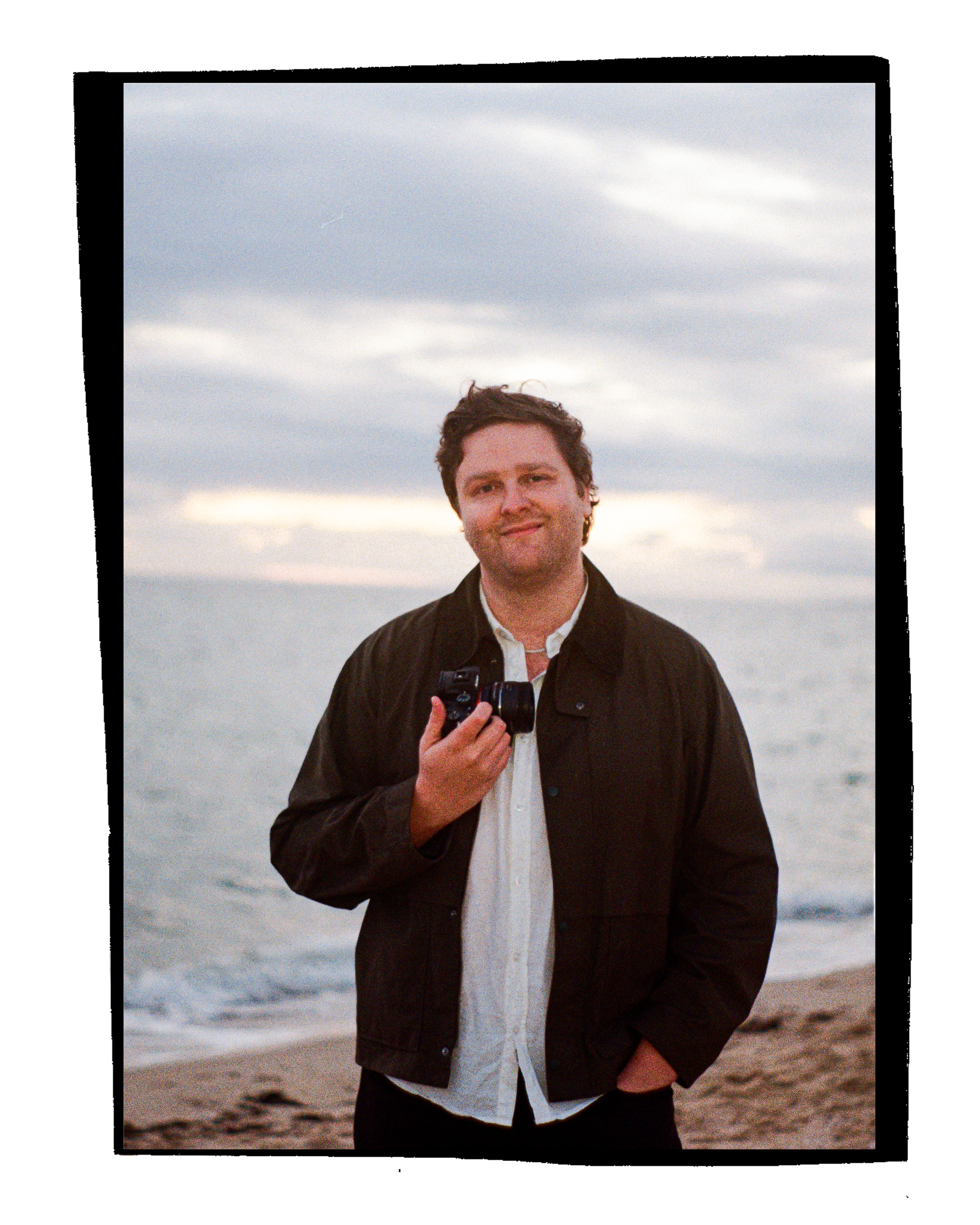 A man standing on a beach holding a camera, wearing a black jacket and white shirt, with ocean and cloudy sky behind him in the evening.