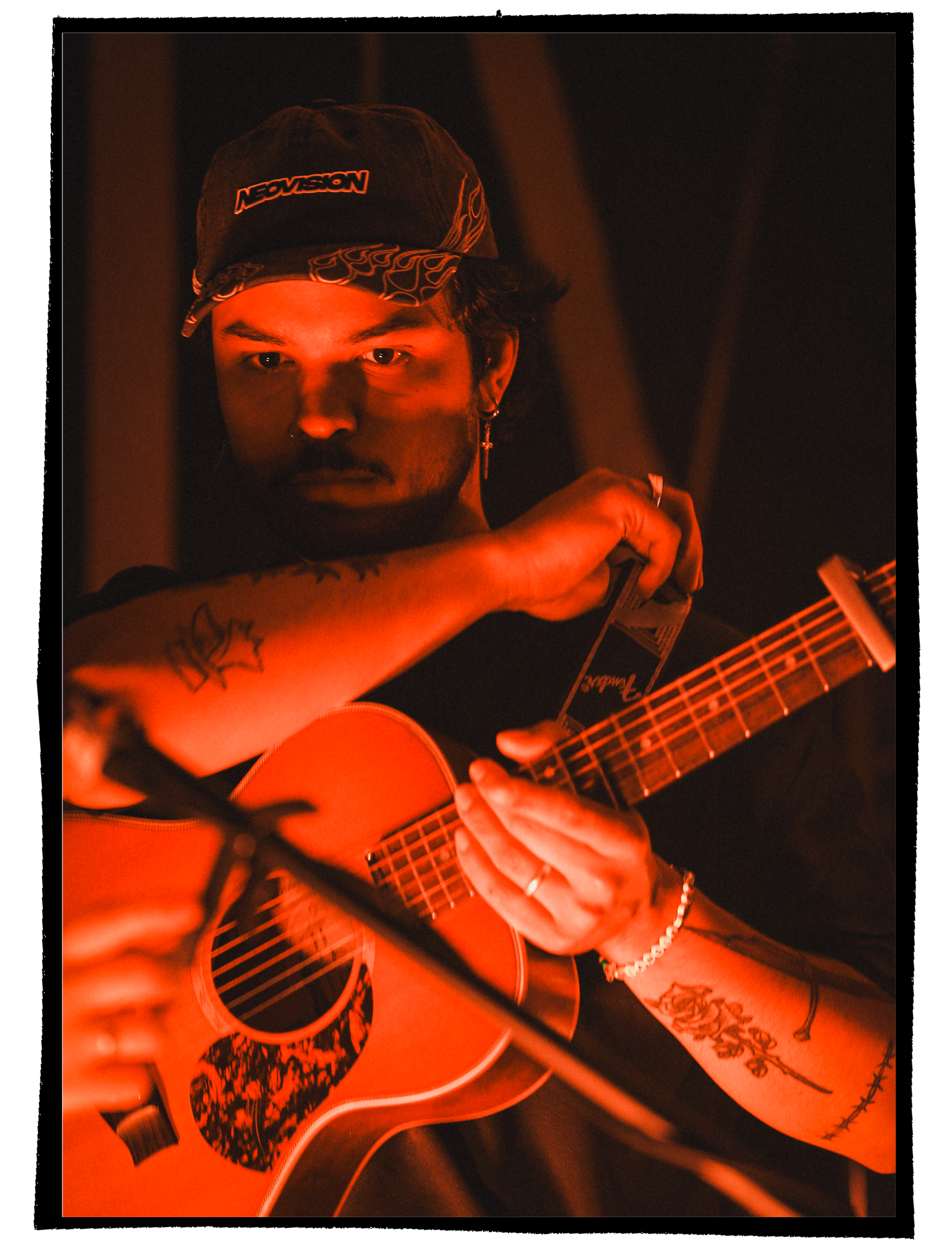 A person with tattoos playing an acoustic guitar in a dimly lit setting with orange-red lighting, wearing a black cap with the word 'NEOVISION' on it, and a mini cross earrings.