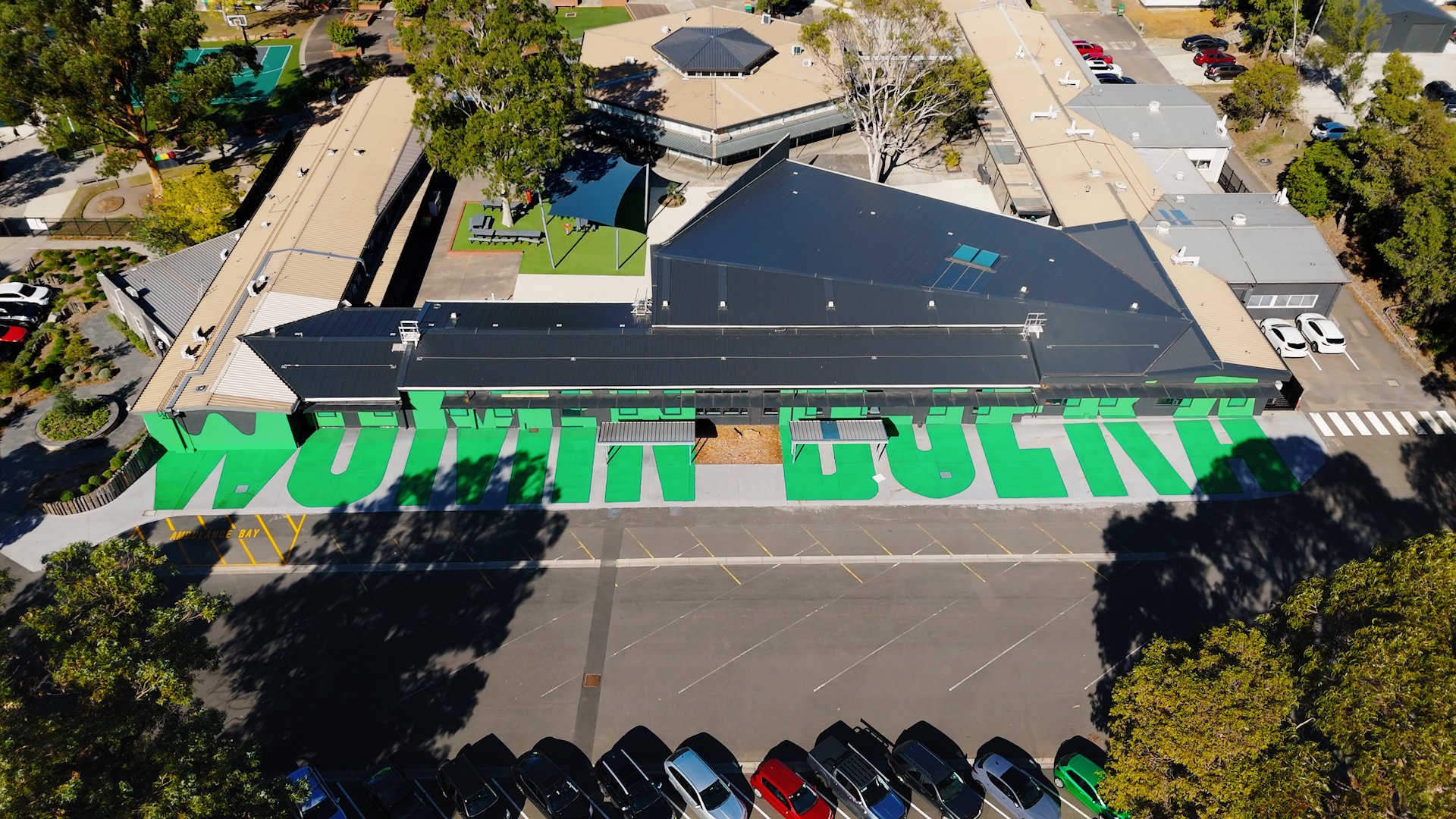 An aerial view of a building with a large green mural that reads 'HUMANS BEING' on the side, surrounded by trees, parking lots, and recreational areas.