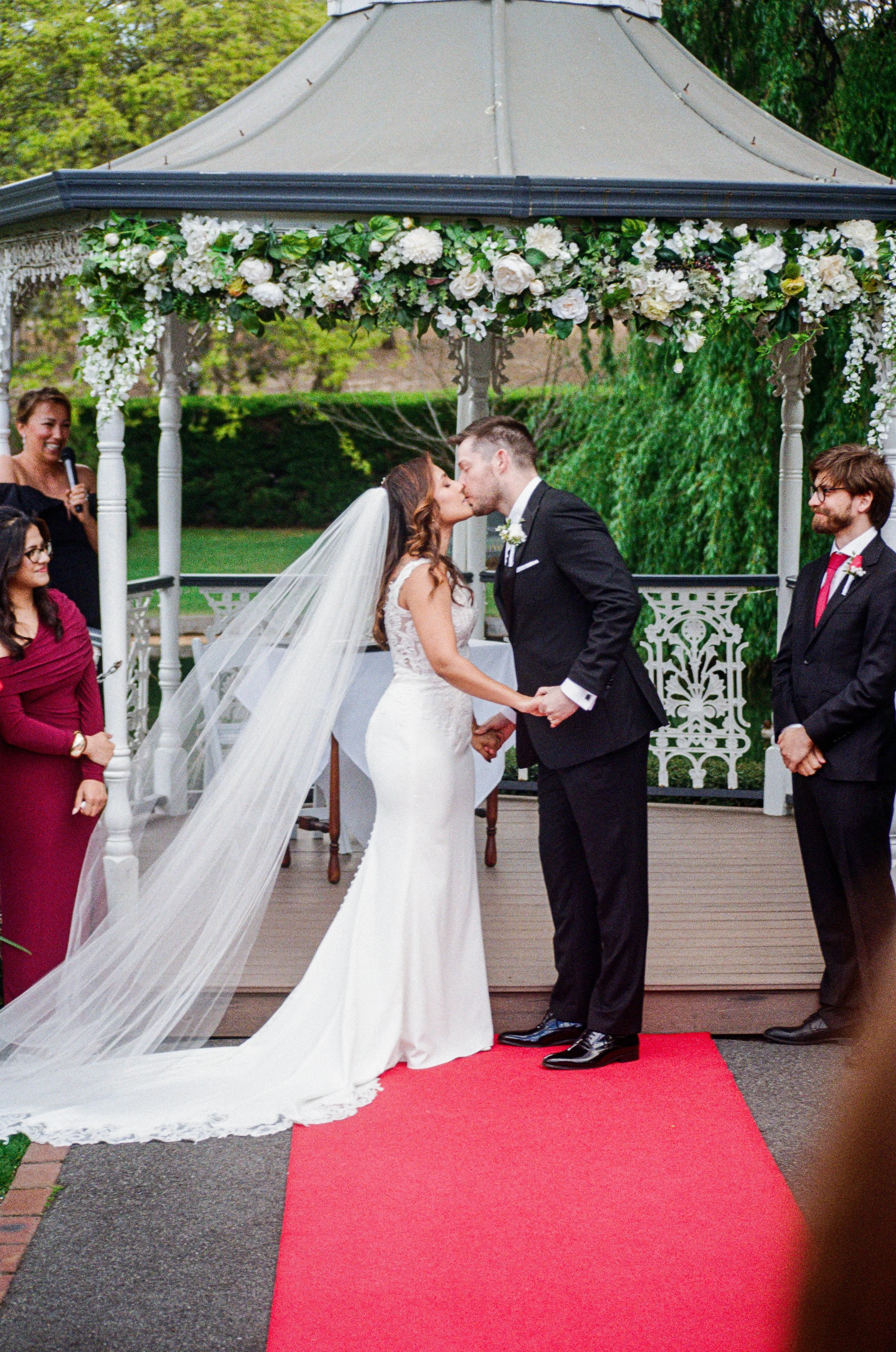A wedding ceremony with a bride and groom kissing under a decorated outdoor gazebo. The couple holds hands on a red carpet, and guests, including a woman in a red dress and a woman with a microphone, look on.
