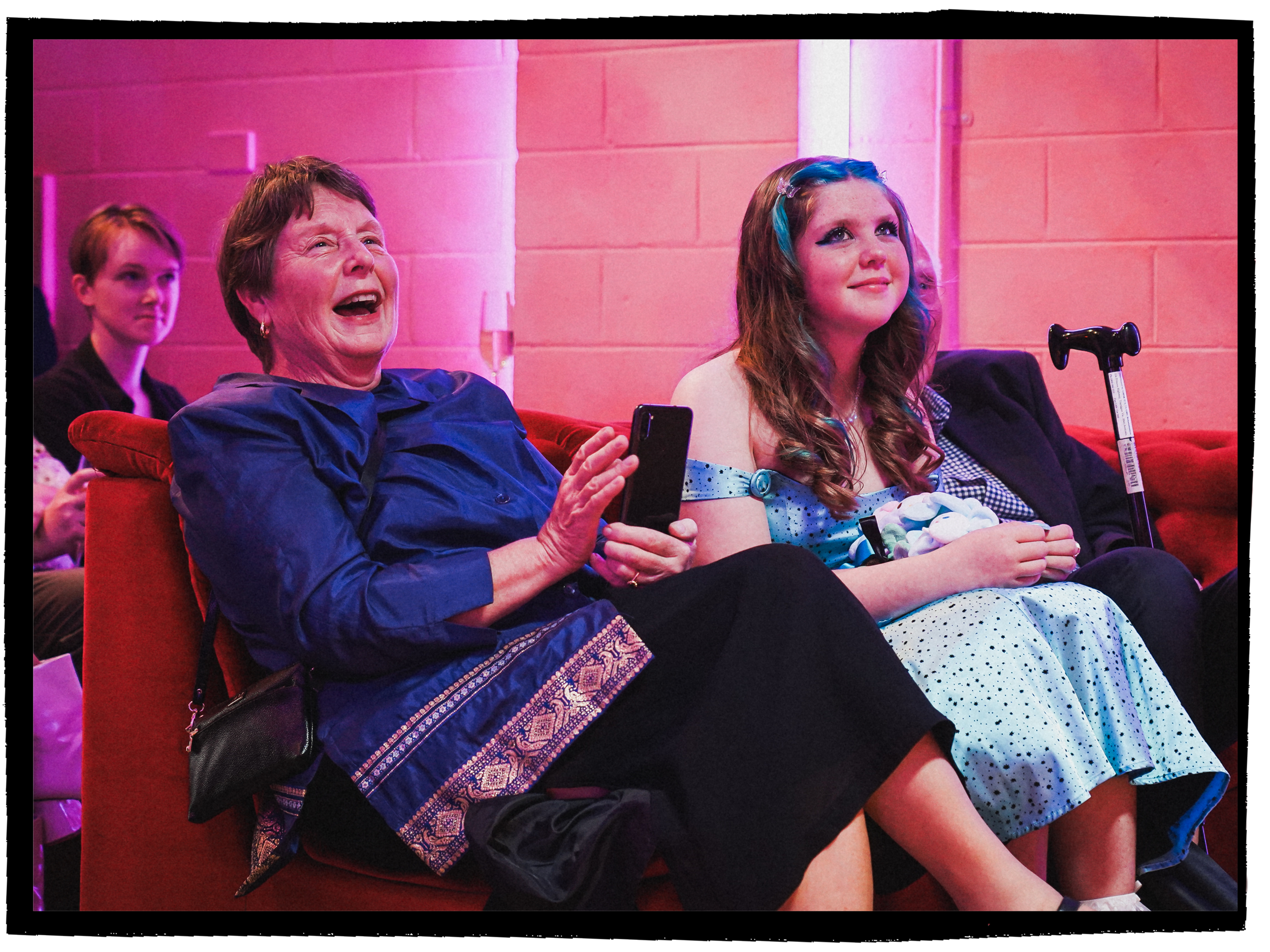 Grandmother and daughter sitting front row at a colourful wedding, smiling and holding phones, with pink lighting and a pink brick wall background.