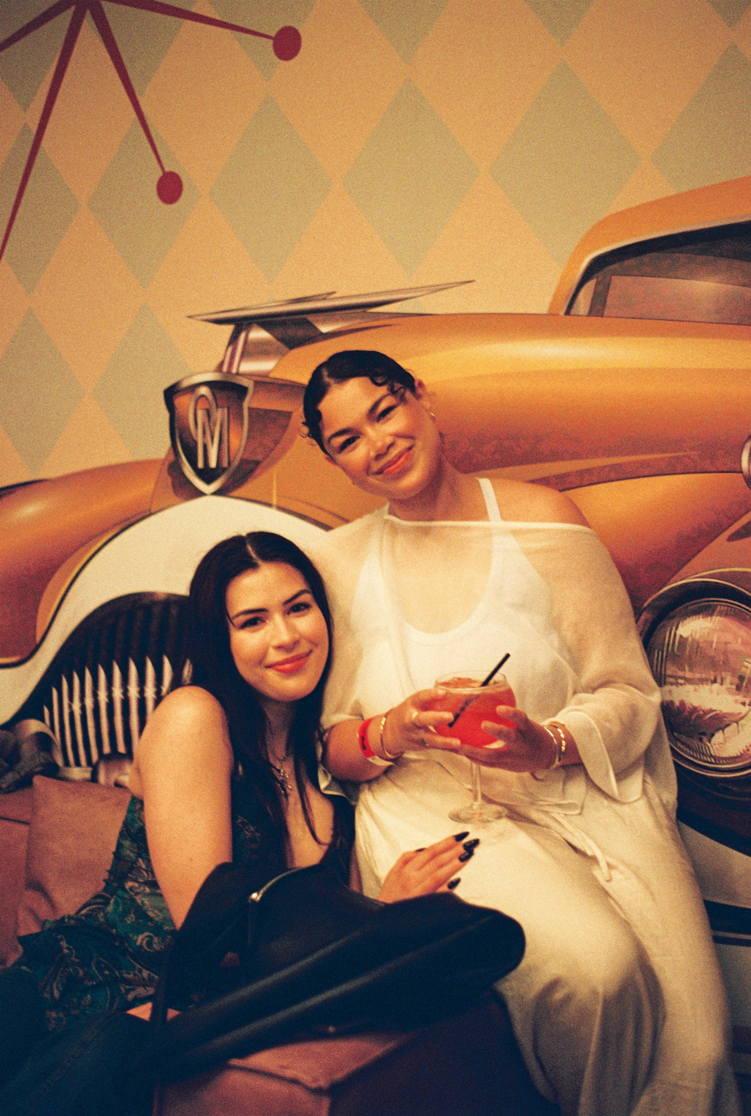 Two women posing for a photo in a colorful retro-themed setting. One woman with long dark hair is sitting, and another woman with short dark hair and a white outfit is standing, holding a glass with a pink drink and a straw. They are smiling in front