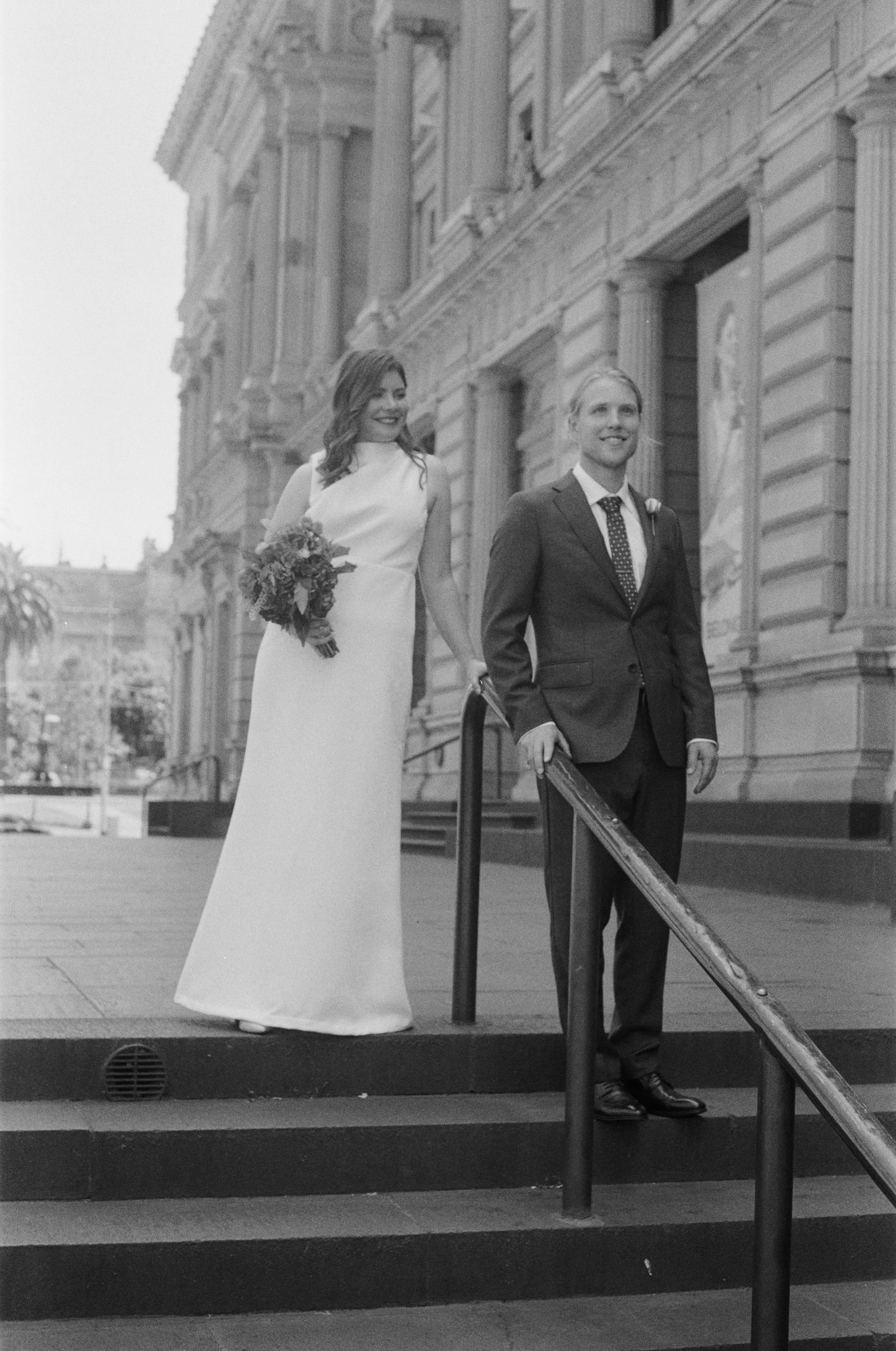A black and white photo of a bride and groom standing on an outdoor staircase in front of a large historic building with ornate architectural details. The bride is holding a bouquet of flowers and wearing a white wedding dress, while the groom is in a dark suit with a tie, smiling and standing at the top of the stairs.