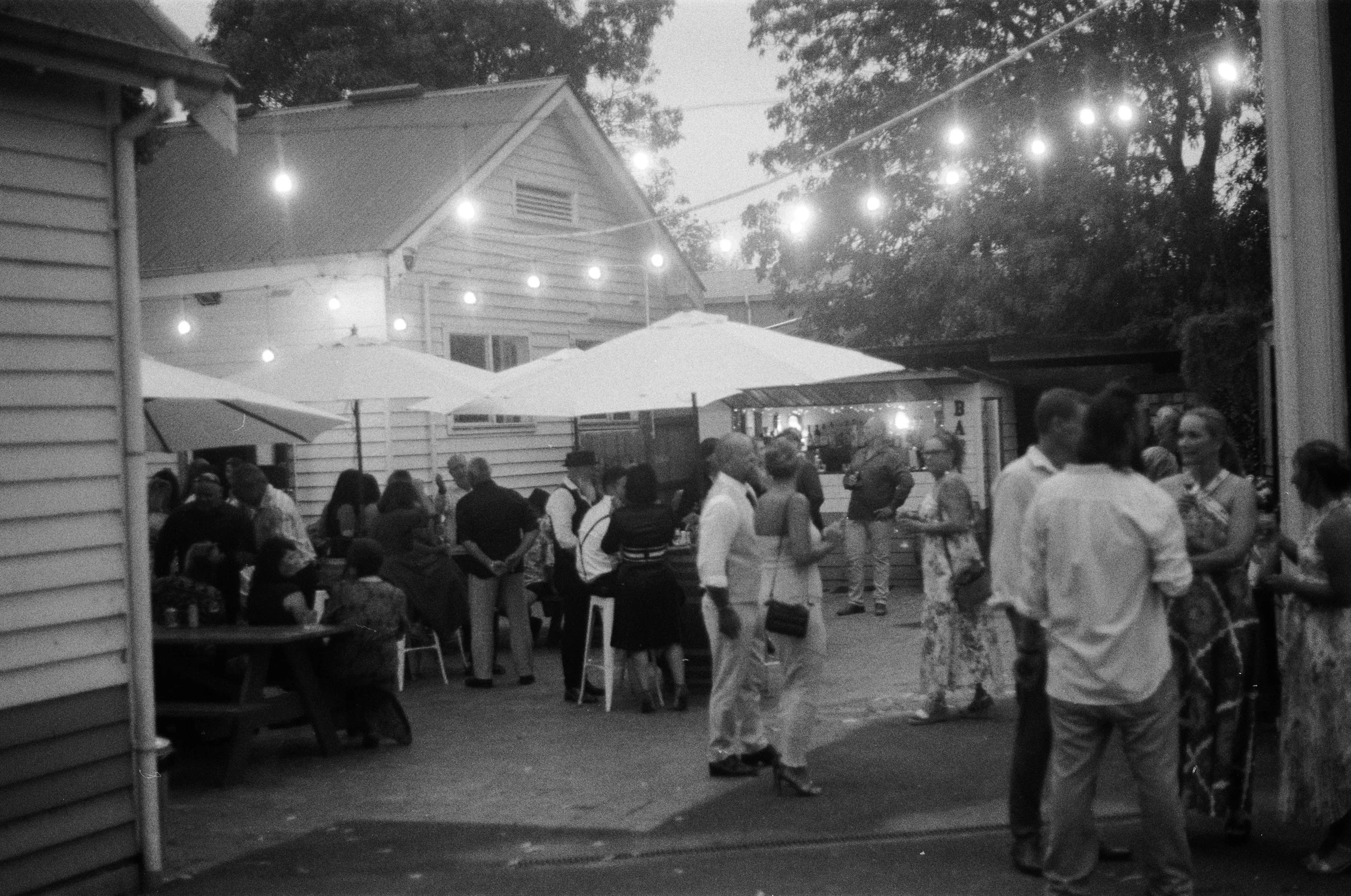 An outdoor gathering scene in the evening with string lights hanging above. Several people are socializing, some sitting and some standing, in a backyard area with umbrellas and a house in the background.