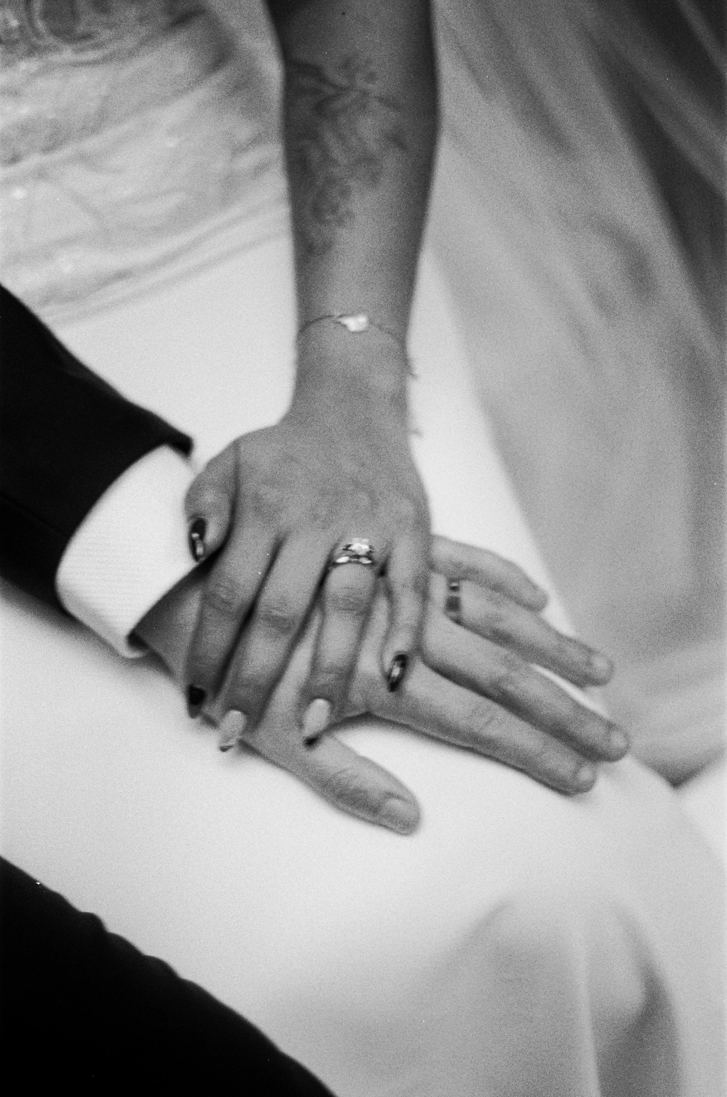 Black and white photo of two hands with rings, one hand resting on the other, on a white surface with parts of clothing visible.