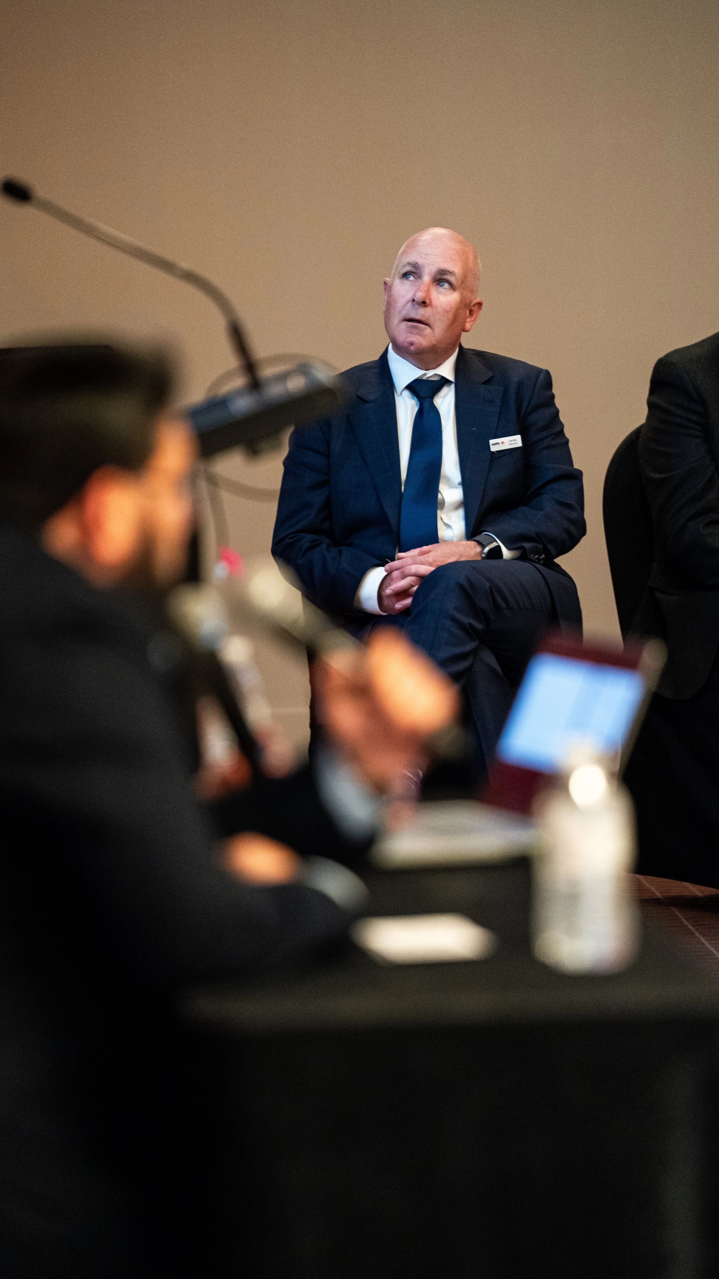 A man in a suit and tie sitting with his hands folded on his lap during a meeting or conference, with a blurred person and recording equipment in the foreground.