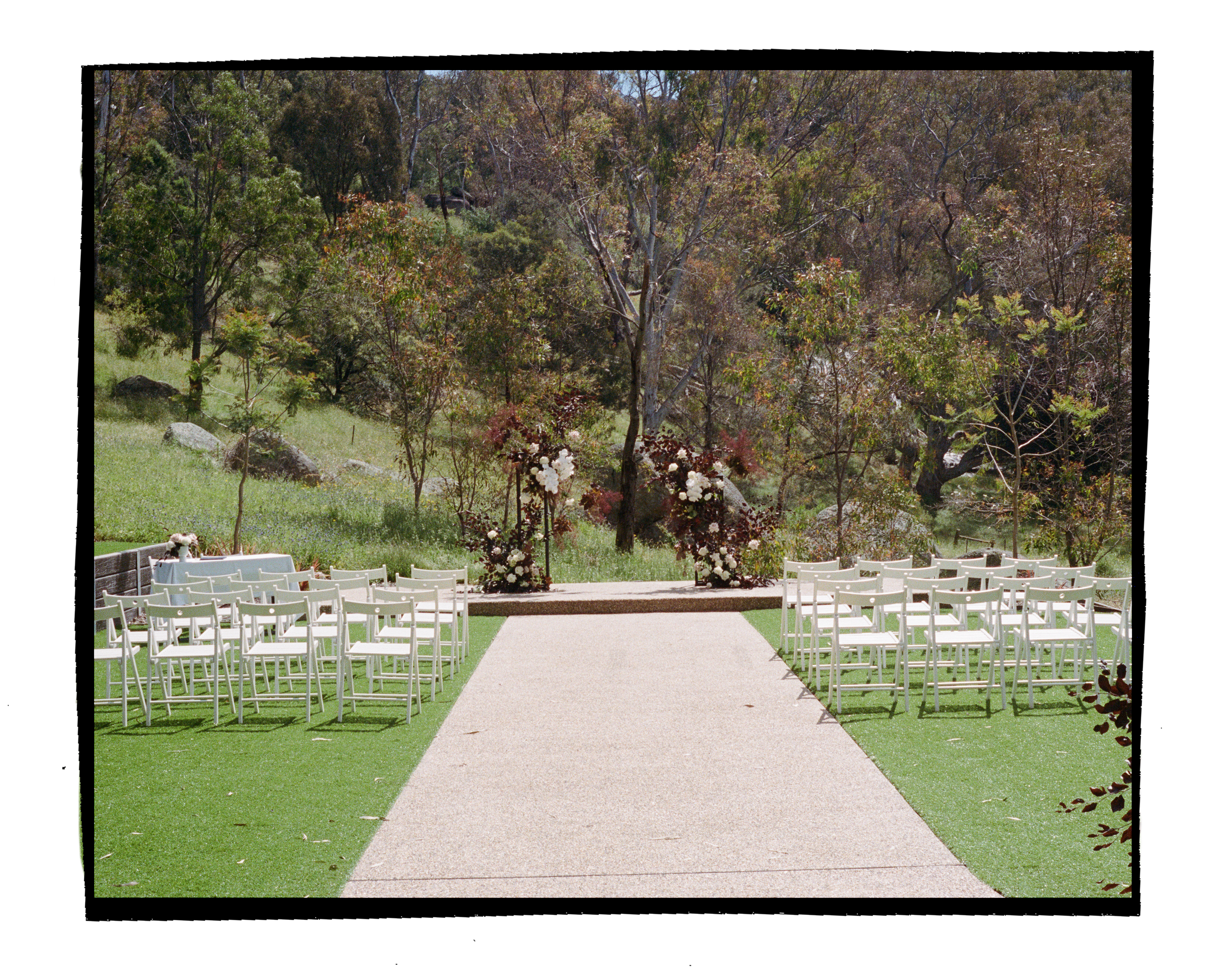 Outdoor wedding ceremony setup with white chairs arranged on either side of a central aisle, flanked by floral arrangements and greenery, with a scenic background of trees and a grassy area.