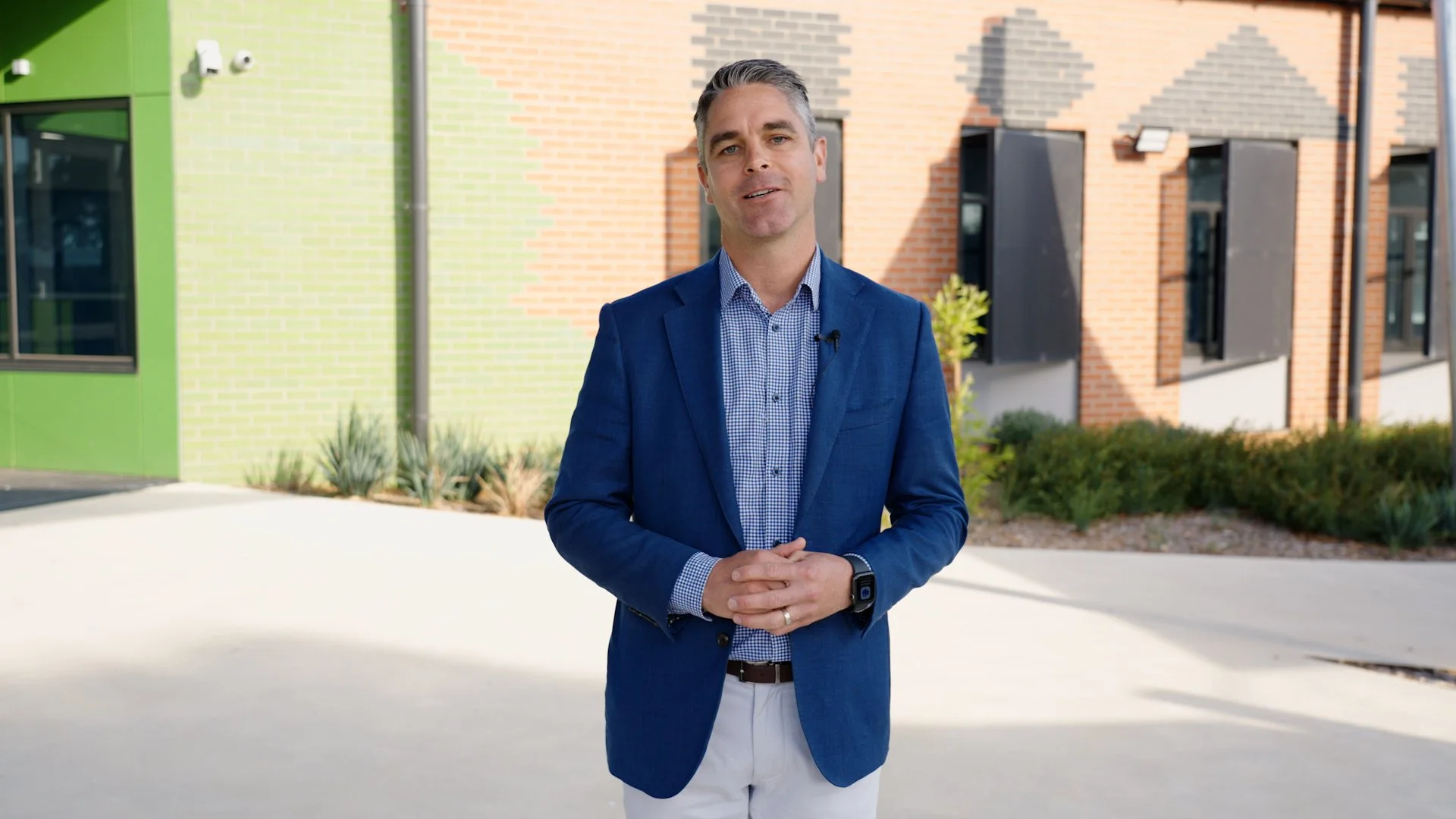 A man wearing a blue blazer and checkered shirt, standing outdoors in front of a modern building with colorful brickwork and windows.