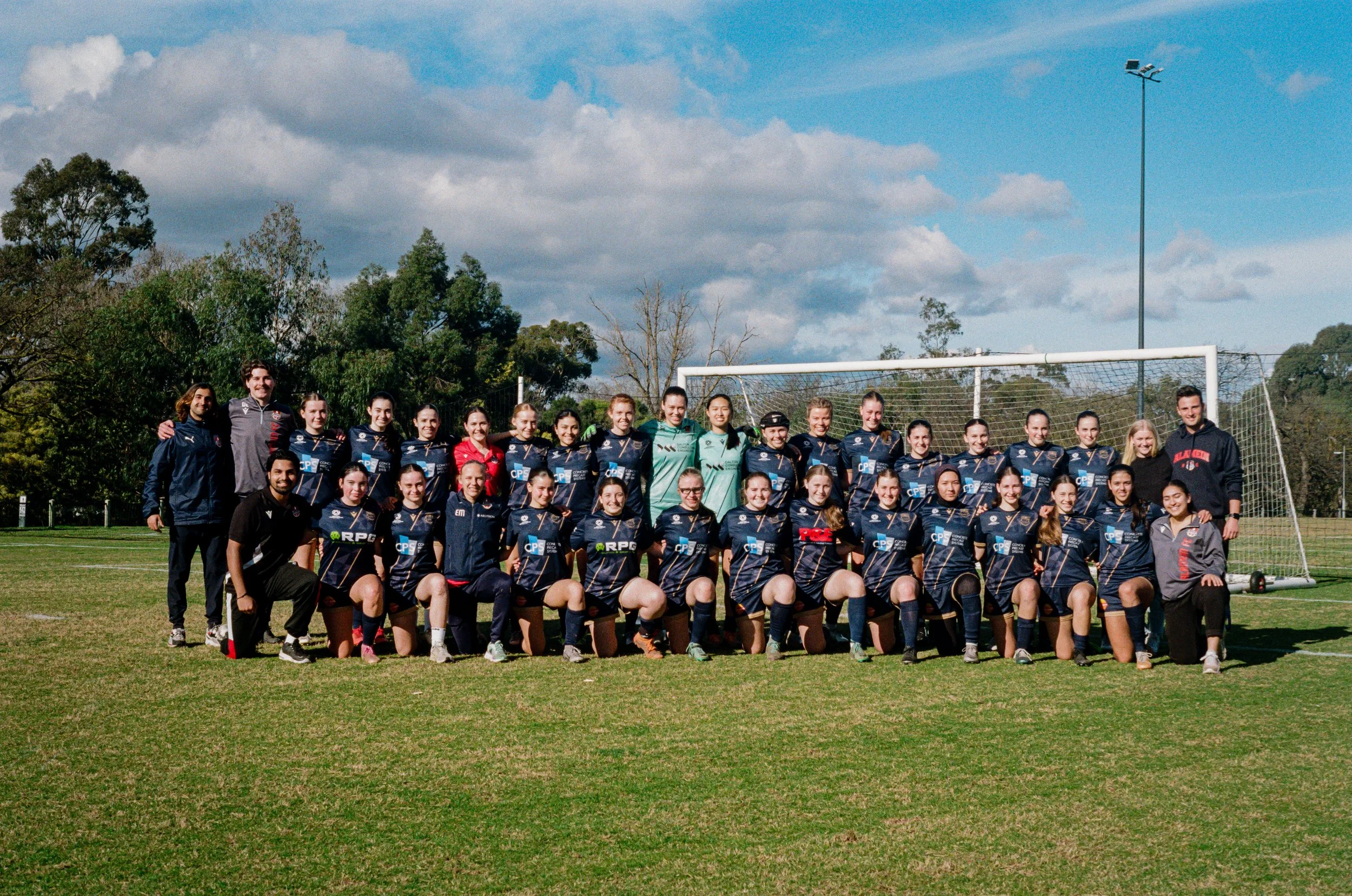 A group of female soccer players and coaches posing for a team photo on a grassy field under a partly cloudy sky, with a soccer goal and trees in the background.