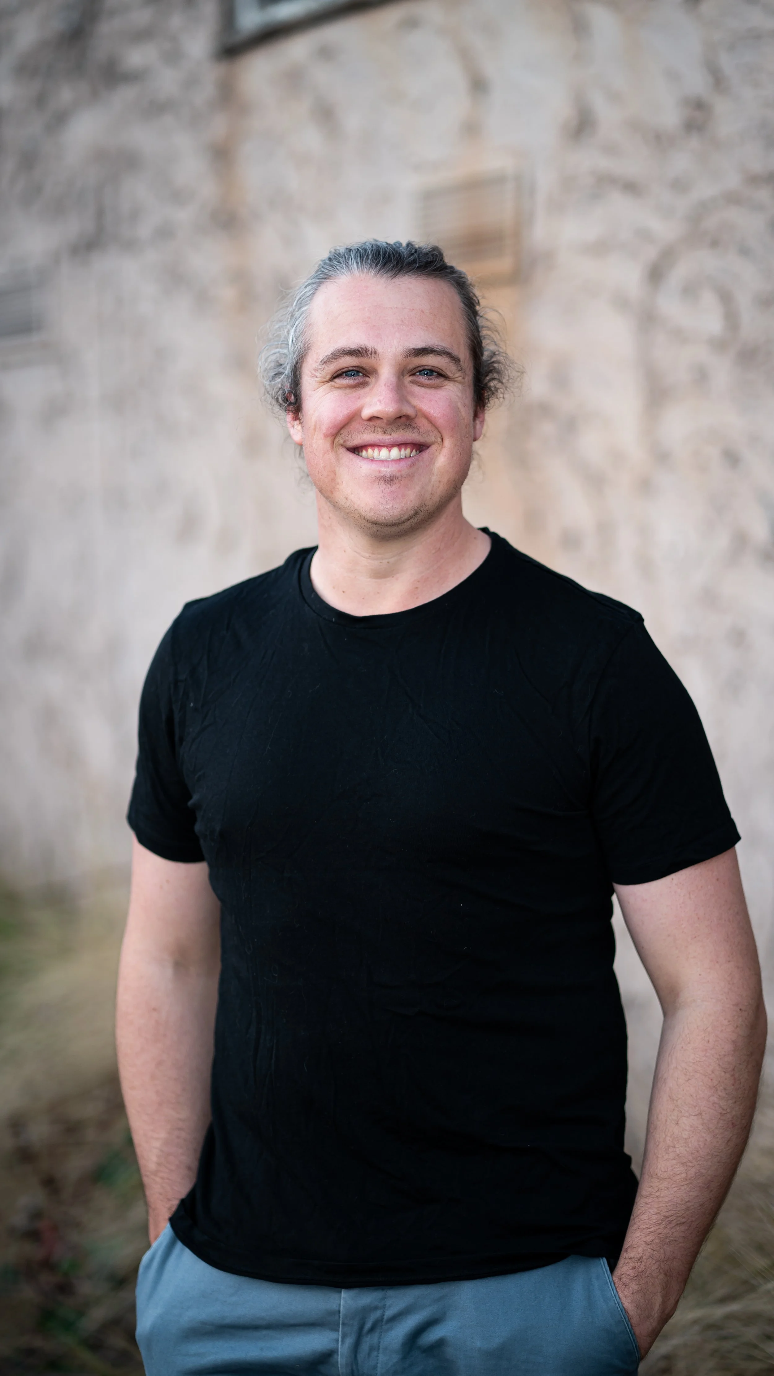 A smiling man with gray hair wearing a black t-shirt and gray pants stands outdoors against a stone wall background.