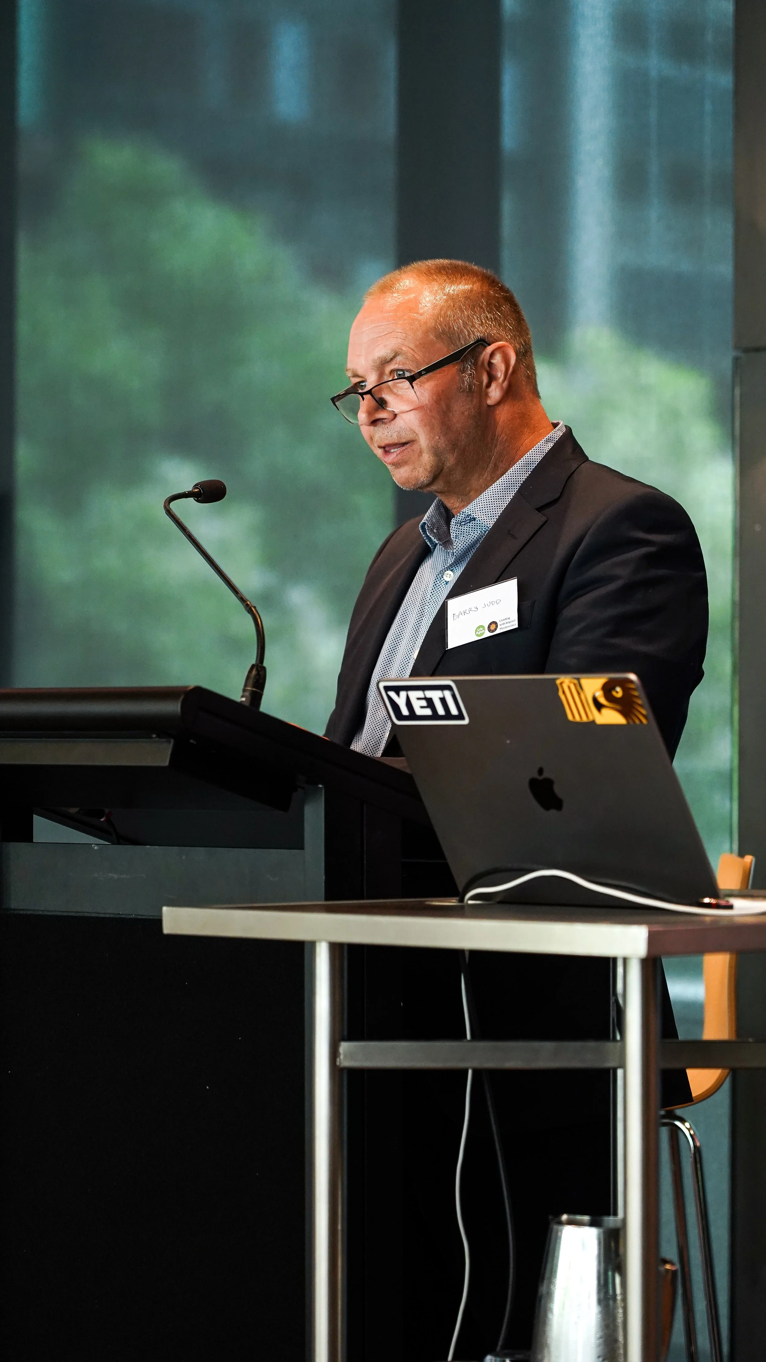 A man in a suit and glasses standing at a podium, speaking into a microphone, with a laptop in front of him labeled YETI and an Apple logo sticker, at a conference or seminar.
