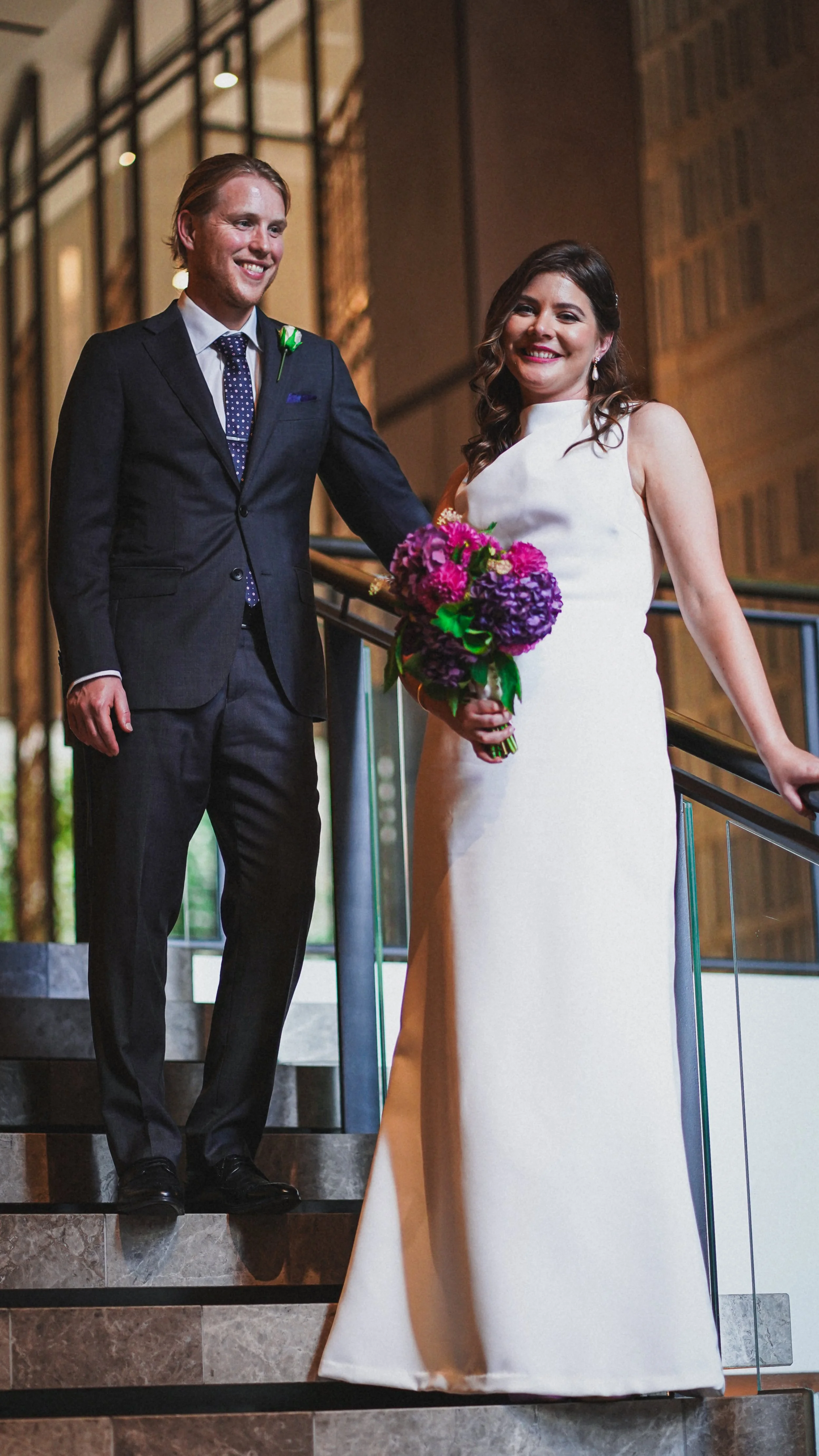 A bride in a white gown and a groom in a dark suit stand on a staircase, smiling and holding a purple flower bouquet.