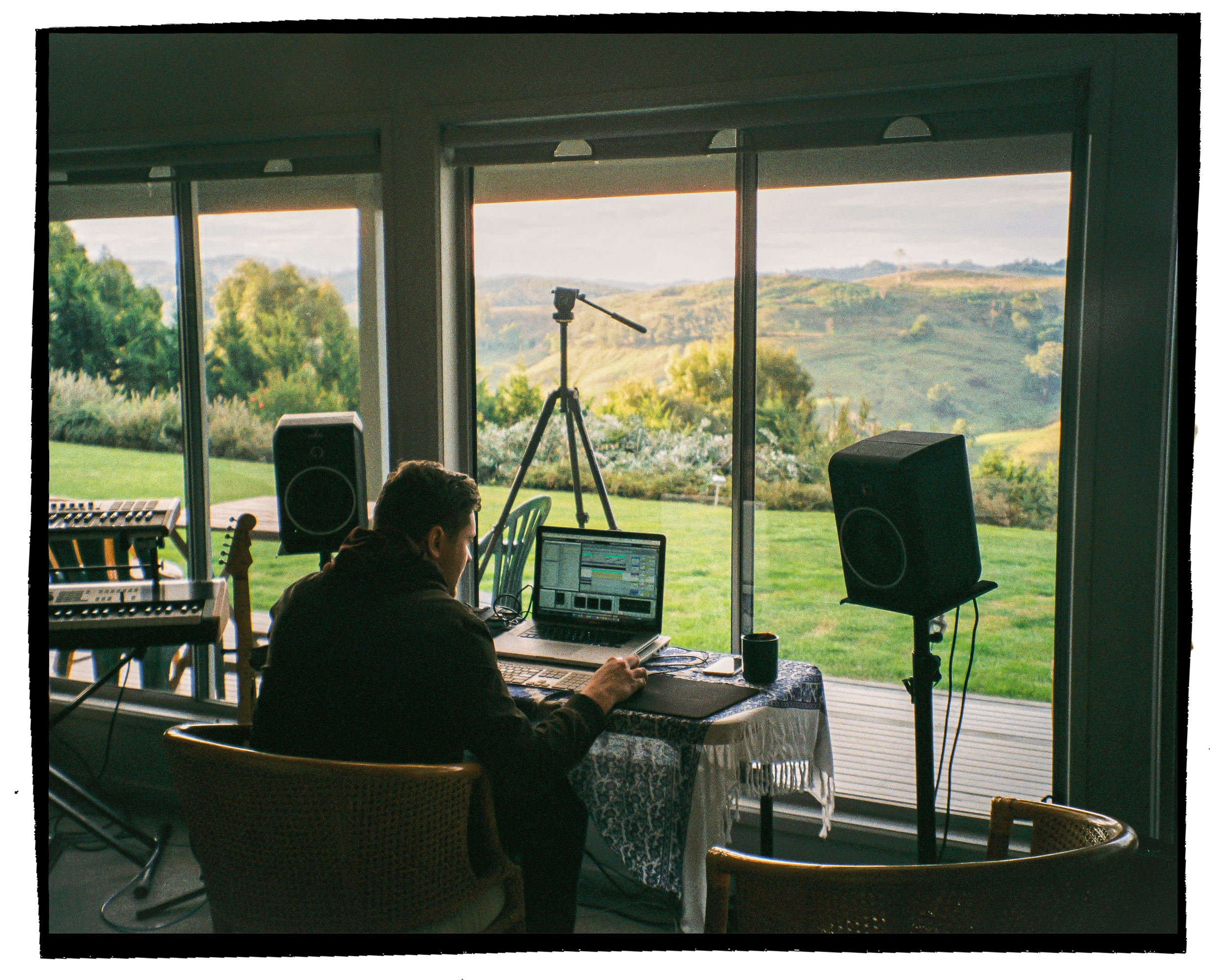 A person sitting at a table working on a laptop, with a scenic view of rolling green hills and trees through large glass windows. Music equipment, including speakers and a keyboard, are visible in the room.