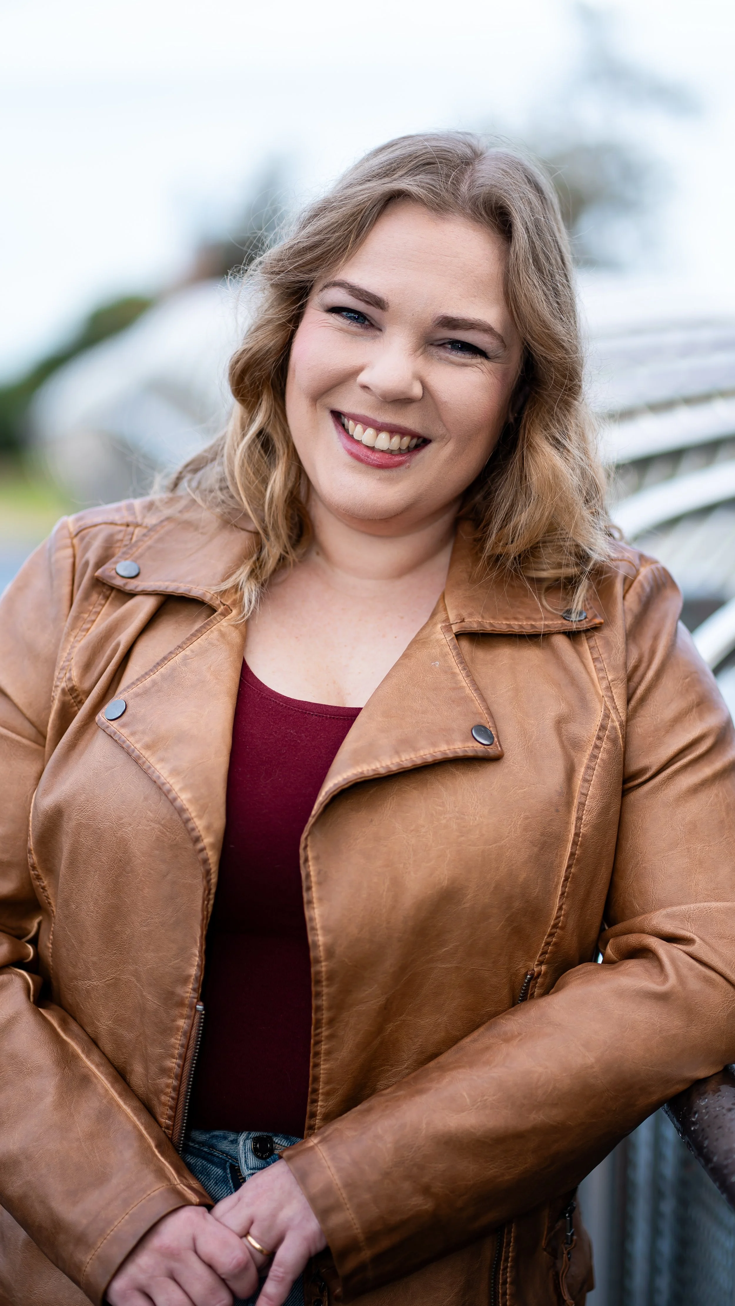 A woman with wavy light brown hair smiling outdoors, wearing a brown leather jacket and a maroon top, with trees and a metal bench in the blurred background.